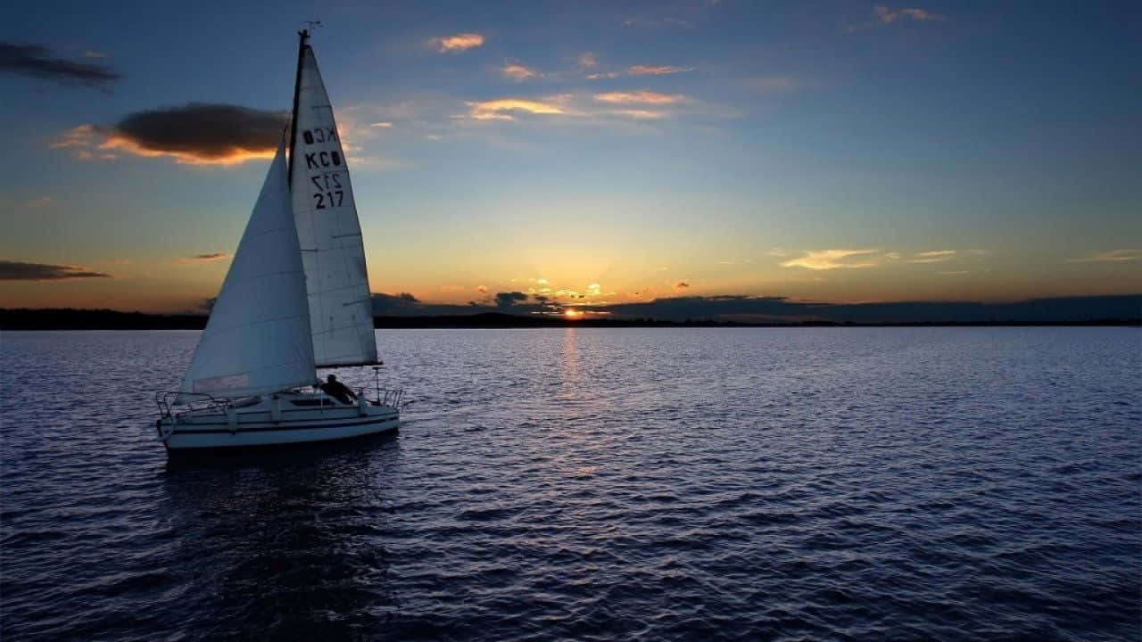 Sailboat 桌布 壁紙 — 一艘帆船在平靜的水面上滑行，夕陽西下，金色光芒映照在波浪上，深藍色的天空。