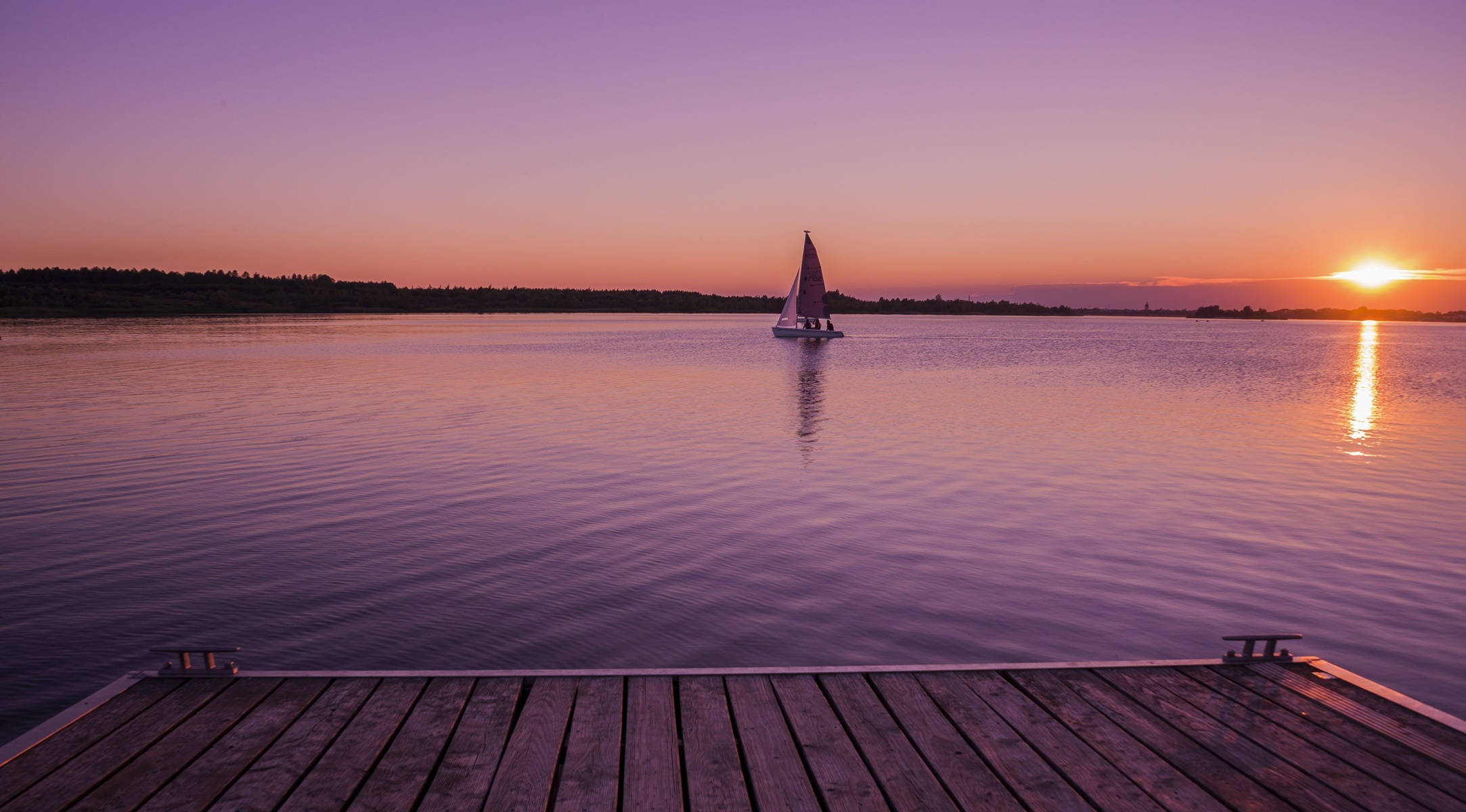 Relaxing Desktop 桌布 壁紙 — 一艘帆船在日落時分滑過寧靜的湖泊，其倒影在平靜的水面上閃爍，從木製碼頭觀看。