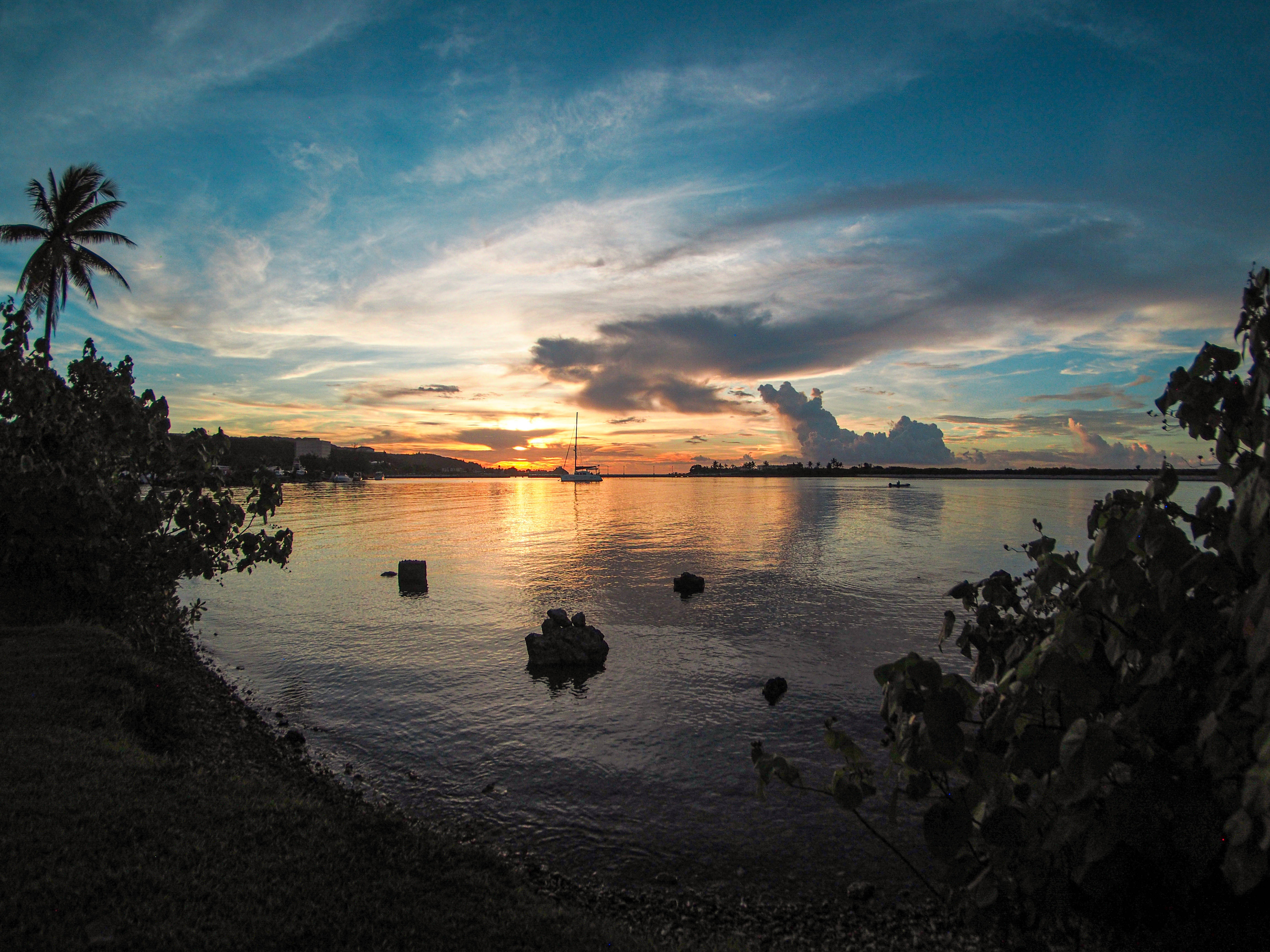 Guam 桌布 壁紙 — 一艘帆船在日落時分滑過平靜的水面， vibrant 的橙色和藍色天空倒映在水面上，並由剪影般的樹葉框住。