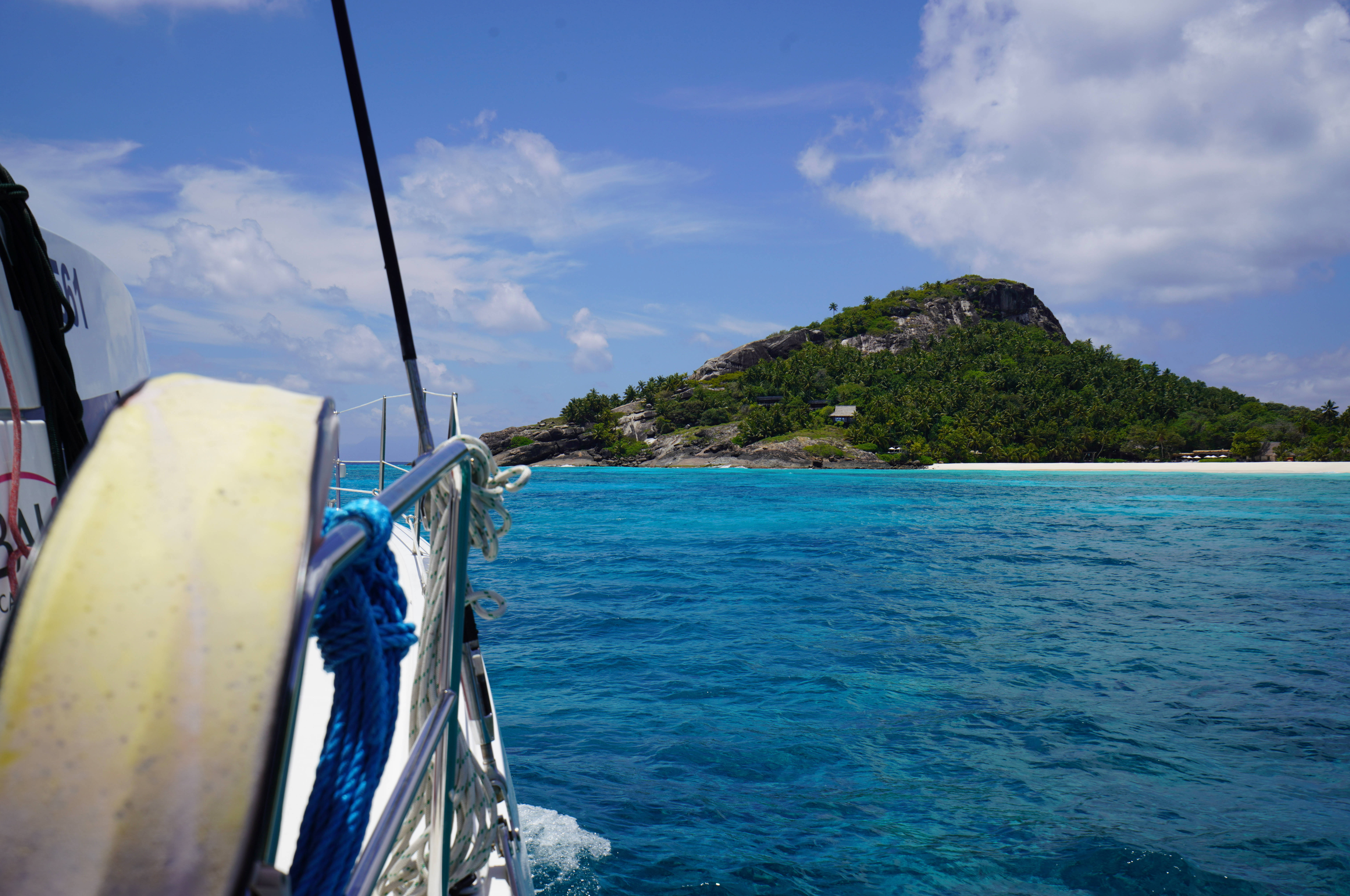 Seychelles 桌布 壁紙 — 一艘帆船在明亮的藍天下，滑過碧綠的海水，駛向一個擁有白色沙灘的茂密綠色島嶼。