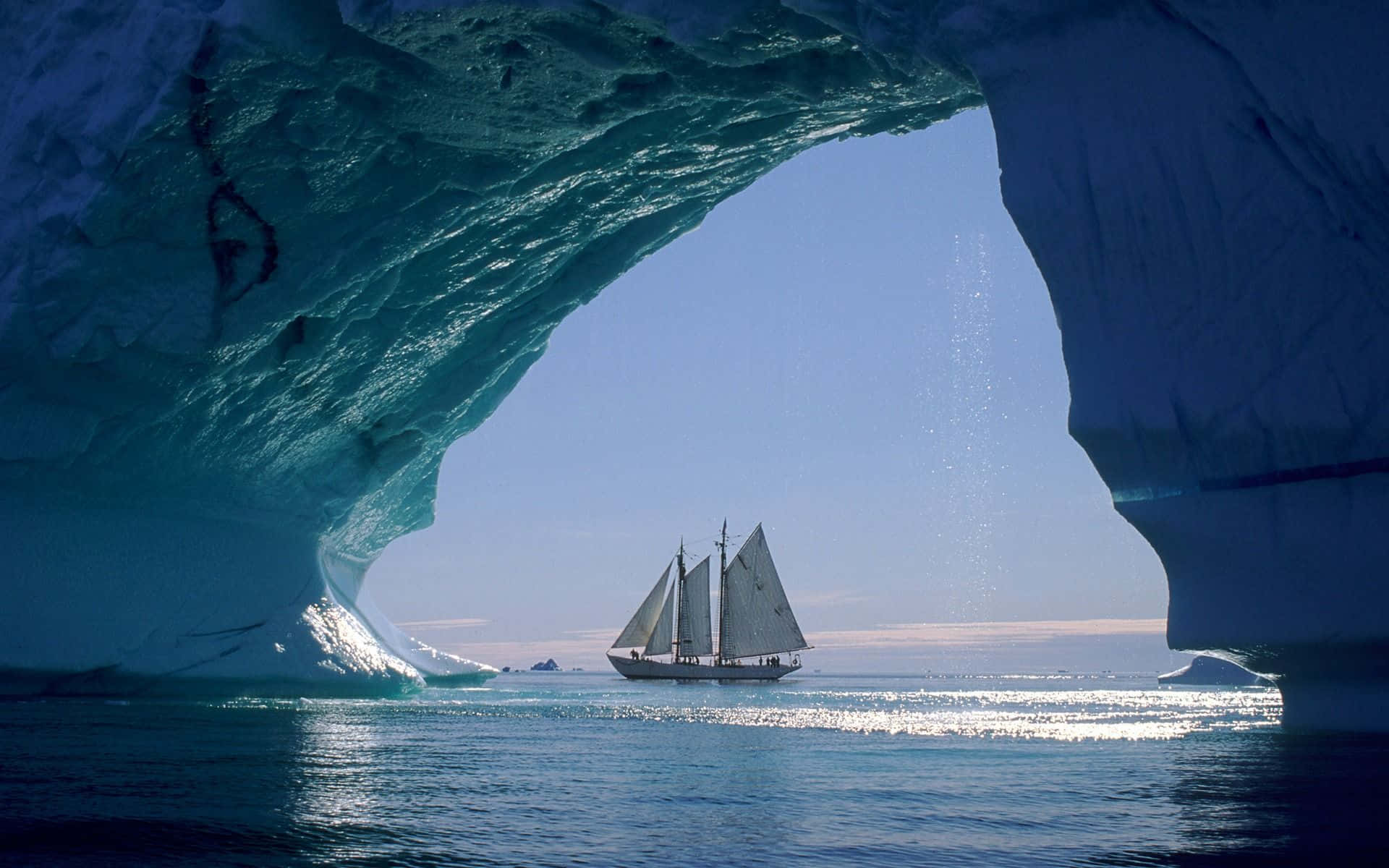 Sailboat 桌布 壁紙 — 一艘帆船在晴朗的天空下滑過極地海洋中發光的藍色冰拱門。