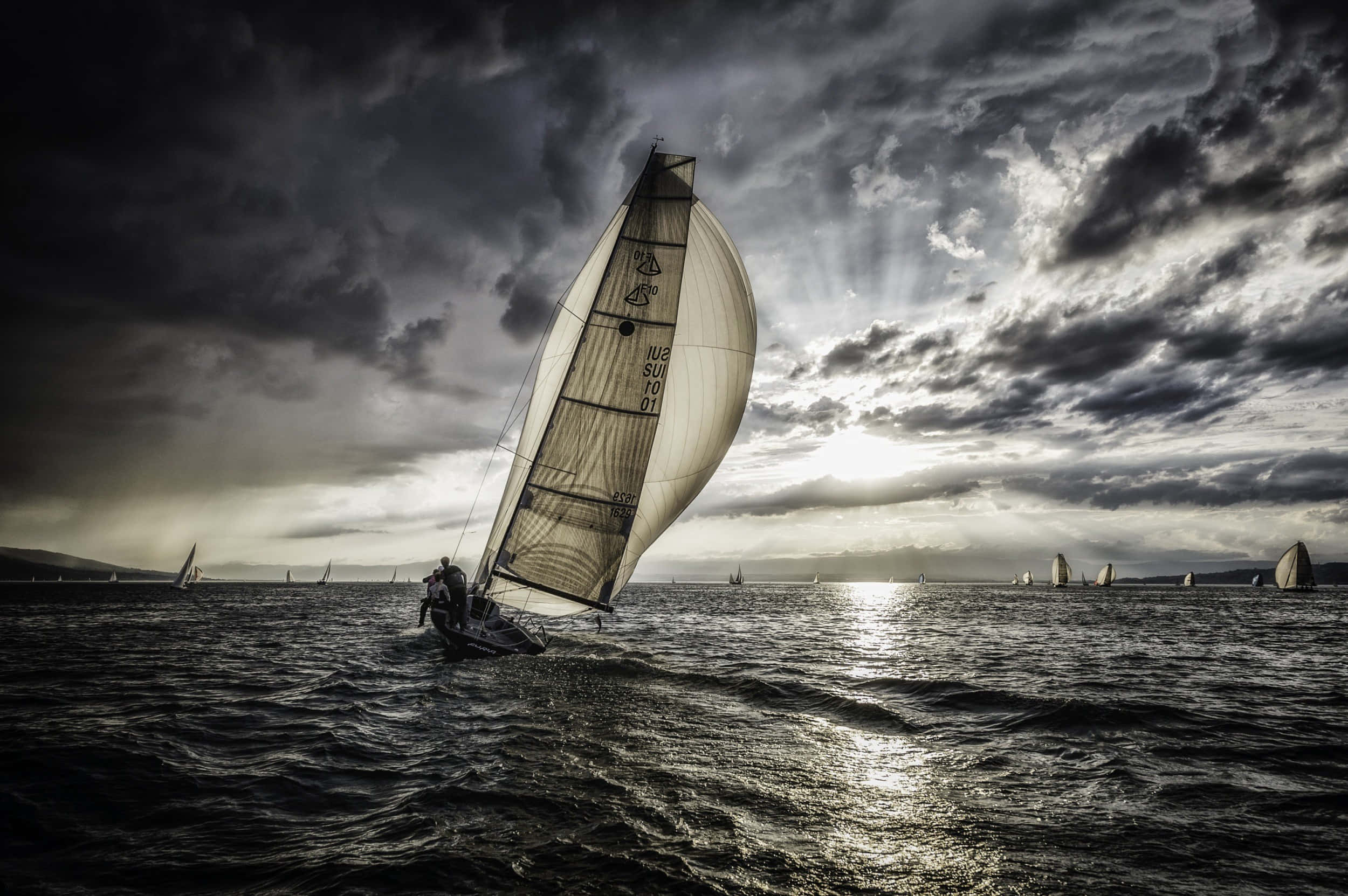 Sailboat 桌布 壁紙 — 一艘帆船在波濤洶湧的水域中破浪前行，天空烏雲密佈，陽光穿透雲層灑下。