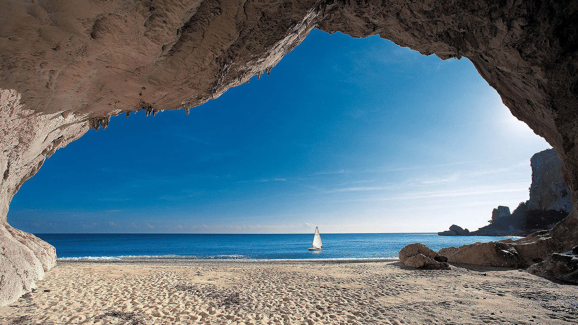 Italian Beach 桌布 壁紙 — 一艘帆船在碧綠色的海面上滑行，透過天然岩石拱門觀看，拱門框著一個陽光明媚的海灘。