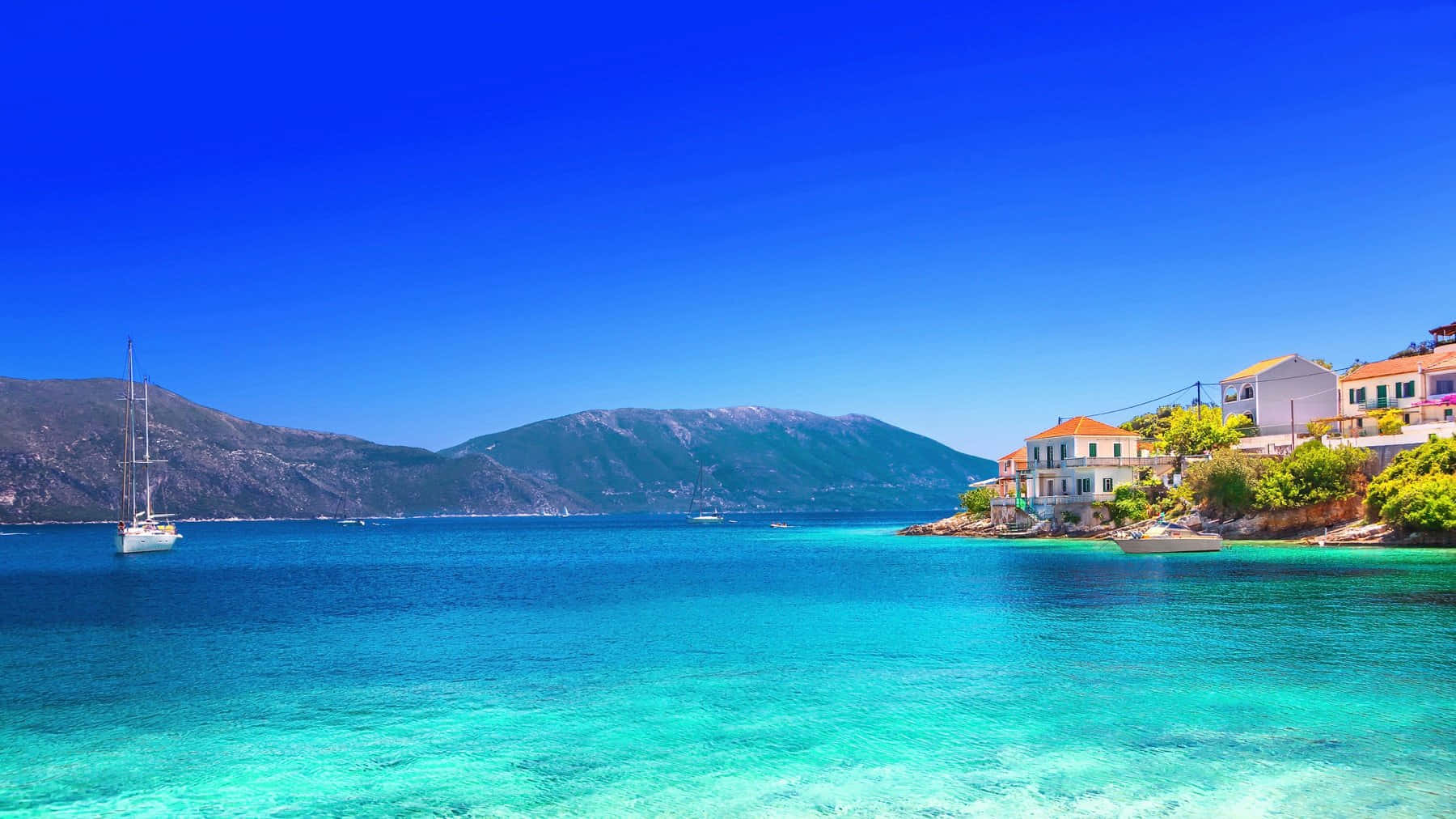Greek Beach 桌布 壁紙 — 一艘帆船漂浮在清澈碧綠的海水上，旁邊是一個懸崖村莊，有白色房屋和陶土屋頂，在充滿活力的藍天下。