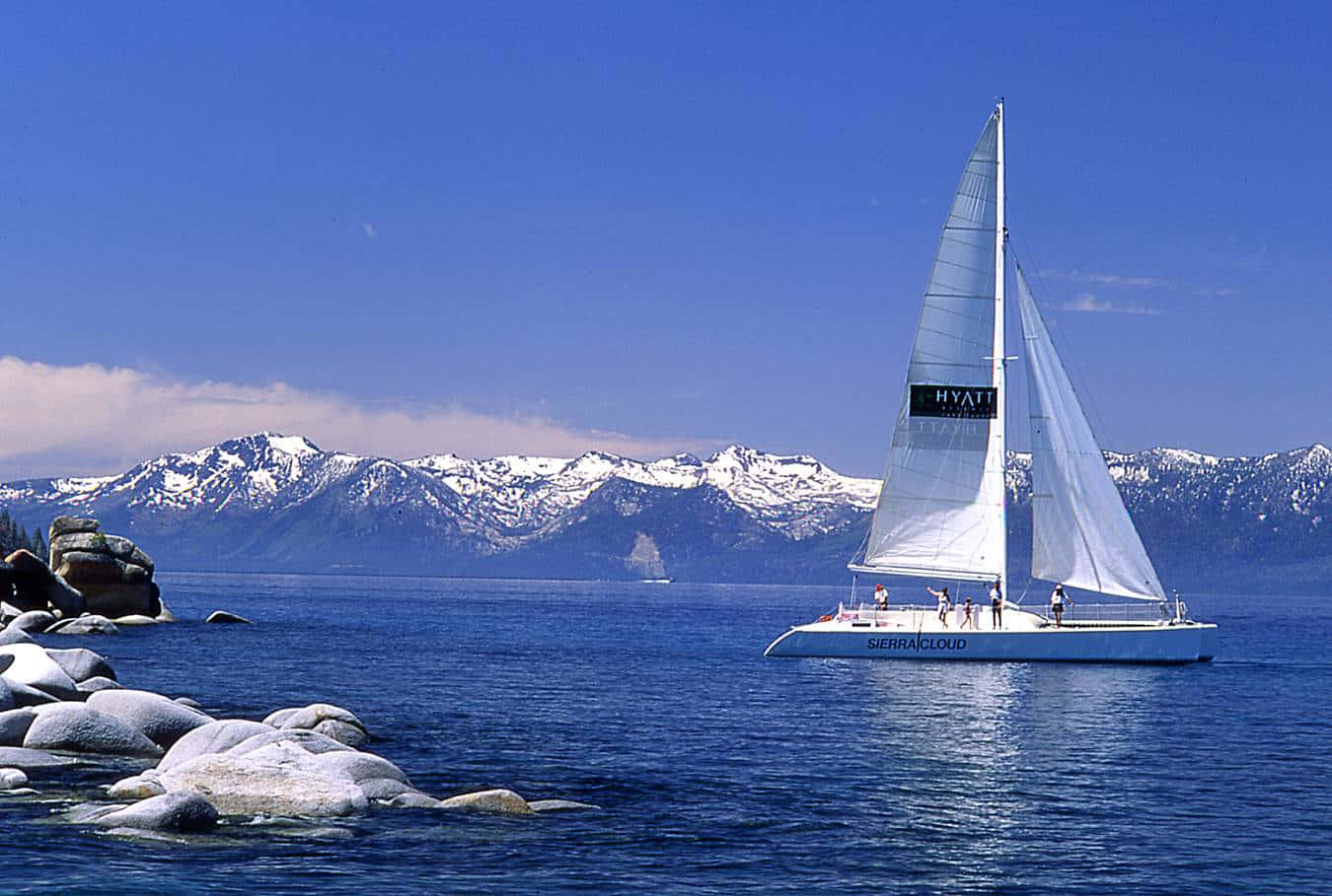Sailboat 桌布 壁紙 — 一艘帶有'HYATT'品牌的白色帆船在清澈藍天下，靠近岩石海岸的深藍色水面上滑行，遠處有積雪覆蓋的山脈。