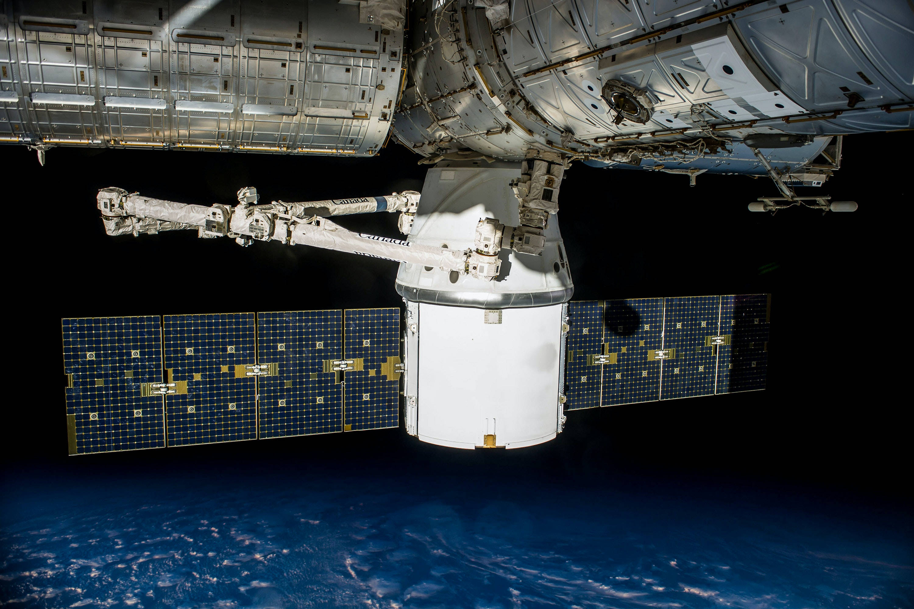NASA 動態圖片 桌布 壁紙 — 一艘帶有太陽能板的太空船從太空站模組延伸出來，面對地球藍色彎曲地平線之上的黑色虛空。