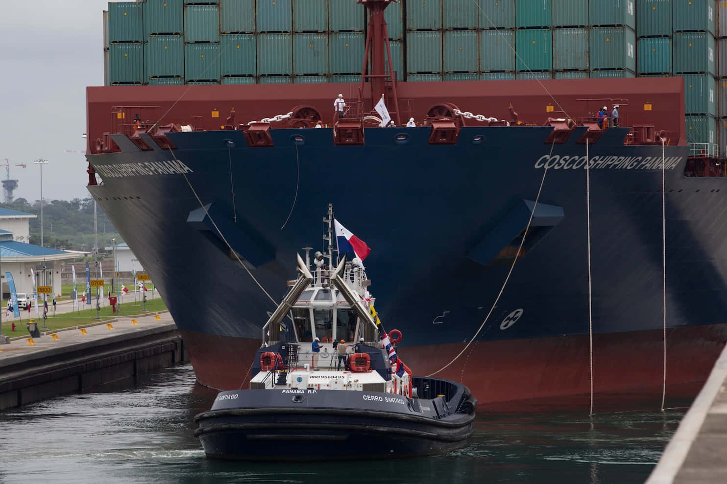 Panama 桌布 壁紙 — 一艘帶有旗幟的小型拖船在運河水閘中，在一艘巨大的藍紅相間貨櫃船旁邊操縱。
