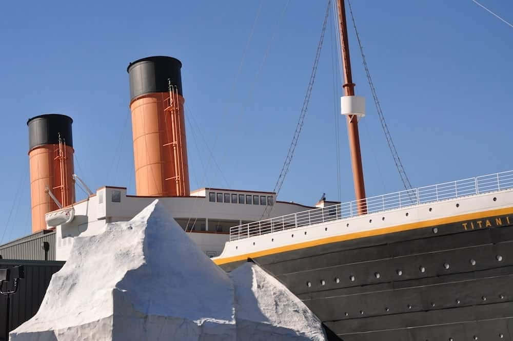 Titanic 桌布 壁紙 — 一艘帶有橙色煙囪和黑色船身的鐵達尼號複製品，在晴朗的藍天下矗立在一個巨大的白色冰山旁邊。