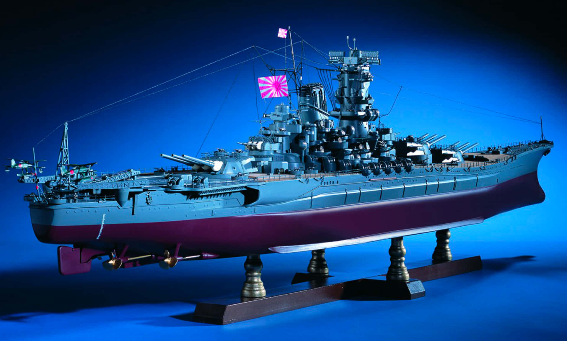 Battleships 桌布 壁紙 — 一艘帶有紅白旗的日本戰艦精細比例模型，矗立在展示架上，背景為深藍色。