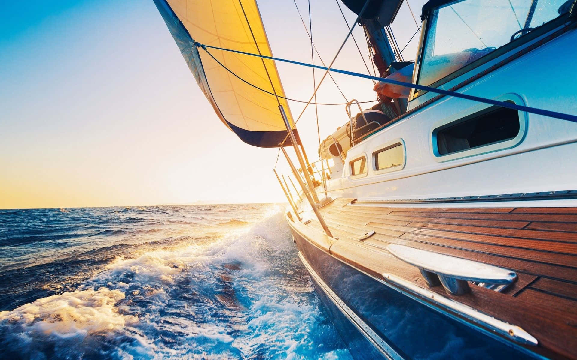 Sailboat 桌布 壁紙 — 一艘帶有黃色帆的帆船在日落時分劃過閃閃發光的藍色海水，將金色光芒灑在木質甲板上。