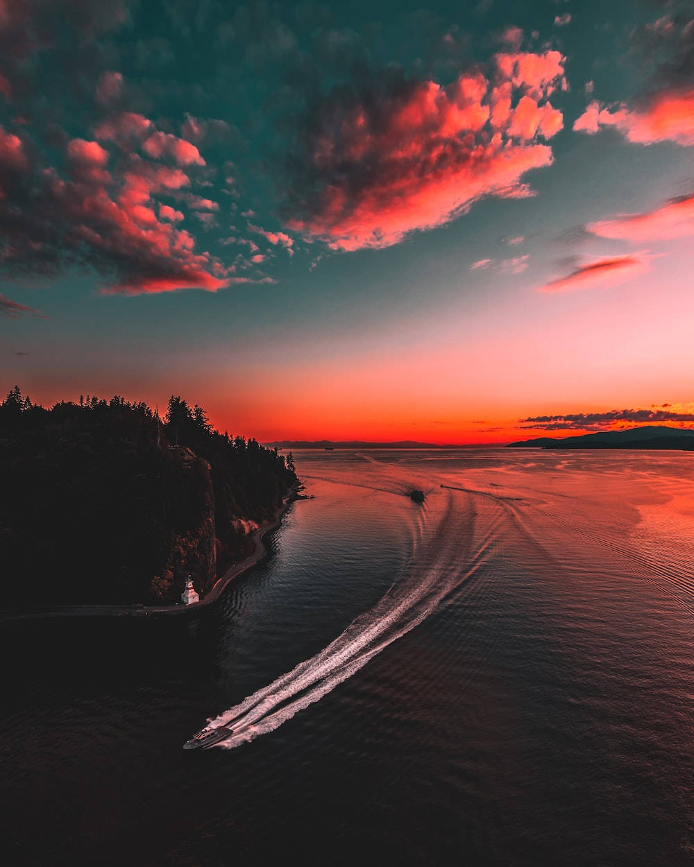 Ocean Sunset 桌布 壁紙 — 一艘快艇在火紅的日落天空下，劃過發光的橙色水面，天空有粉紅色的雲和黑暗的森林懸崖。