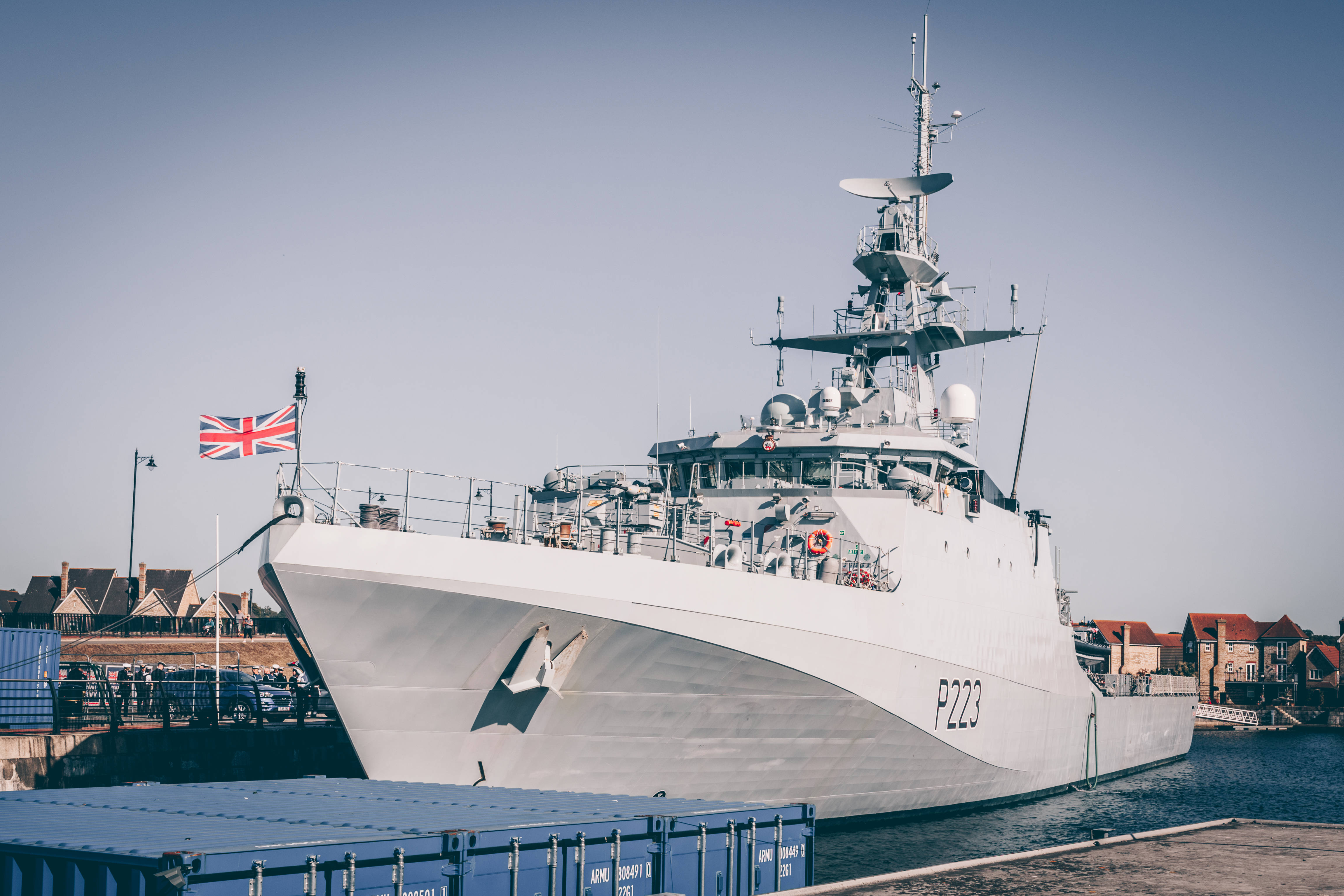 Warship 桌布 壁紙 — 一艘時尚的灰色海軍軍艦，懸掛英國國旗，在平靜的港灣中驕傲地航行，天空湛藍。