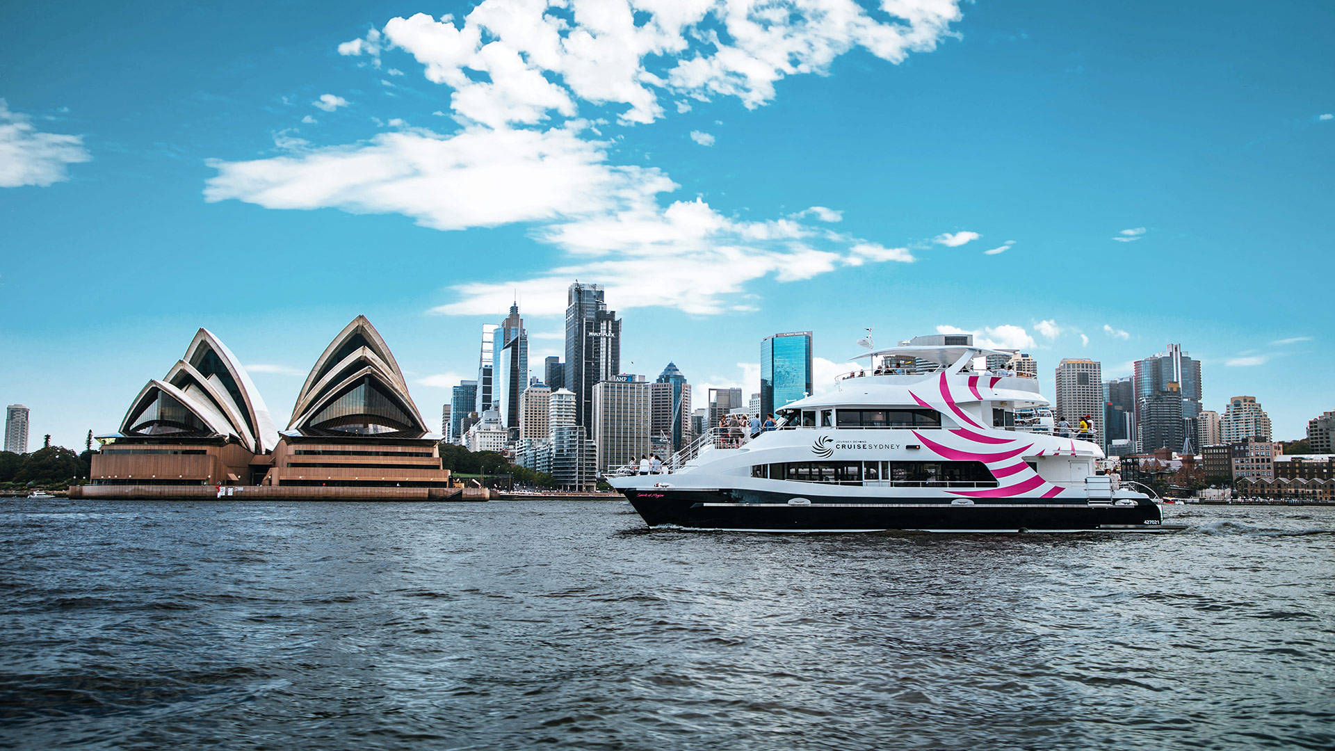 Sydney 桌布 壁紙 — 一艘時尚的白色渡輪帶有粉紅色漩渦，在雪梨港上空滑行，天空湛藍，點綴著蓬鬆的白雲，背景是標誌性的歌劇院和現代天際線。