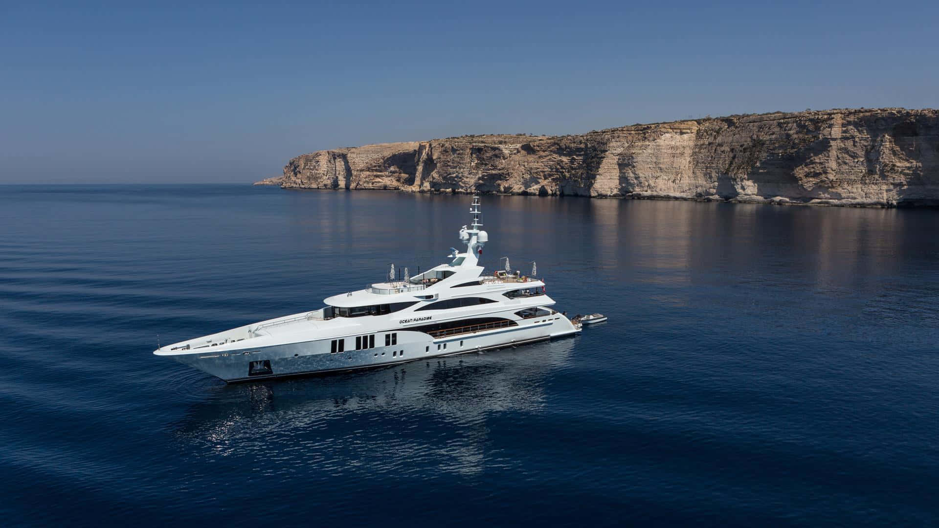 Yacht 桌布 壁紙 — 一艘時尚的白色超級遊艇在晴朗的天空下，在戲劇性的岩石懸崖附近滑過深藍色的海水。