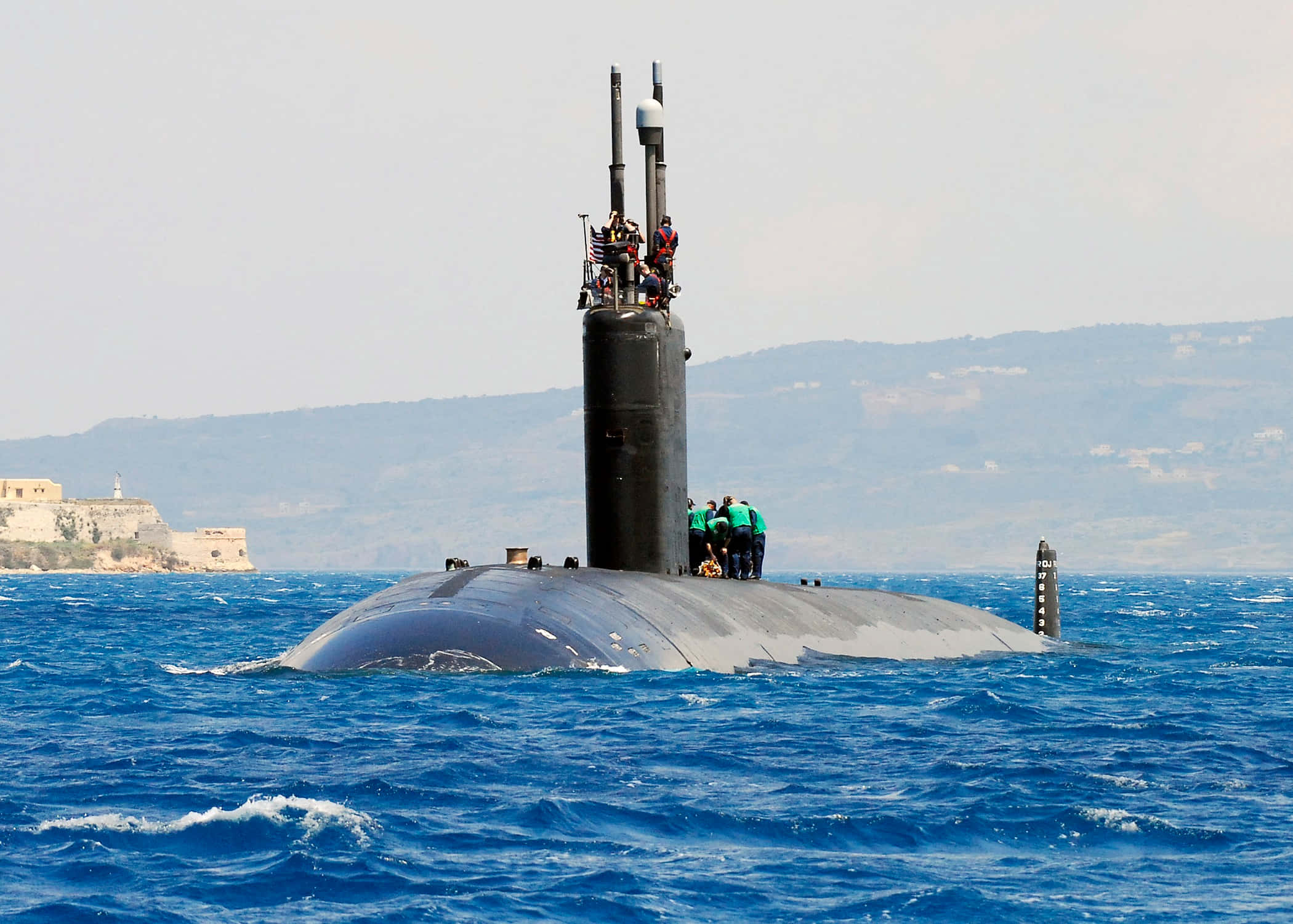 Submarine 桌布 壁紙 — 一艘時尚的黑色潛艇在充滿活力的藍色海洋中浮出水面，船員在甲板上可見，附近有一座遙遠的沿海堡壘。