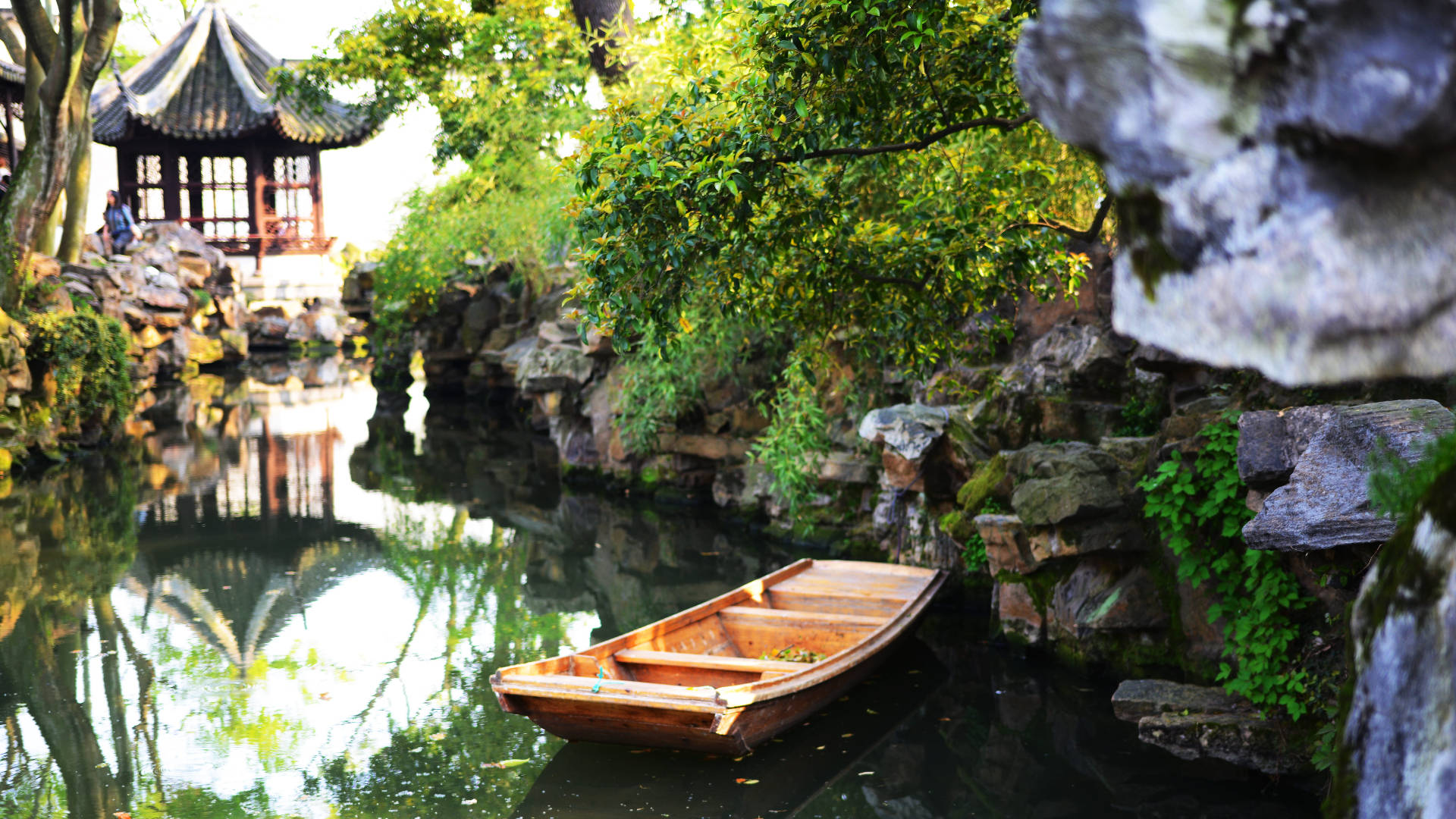 Suzhou 桌布 壁紙 — 一艘木船平靜地停泊在寧靜的運河上，周圍環繞著茂密的綠植和傳統中式亭閣的倒影。