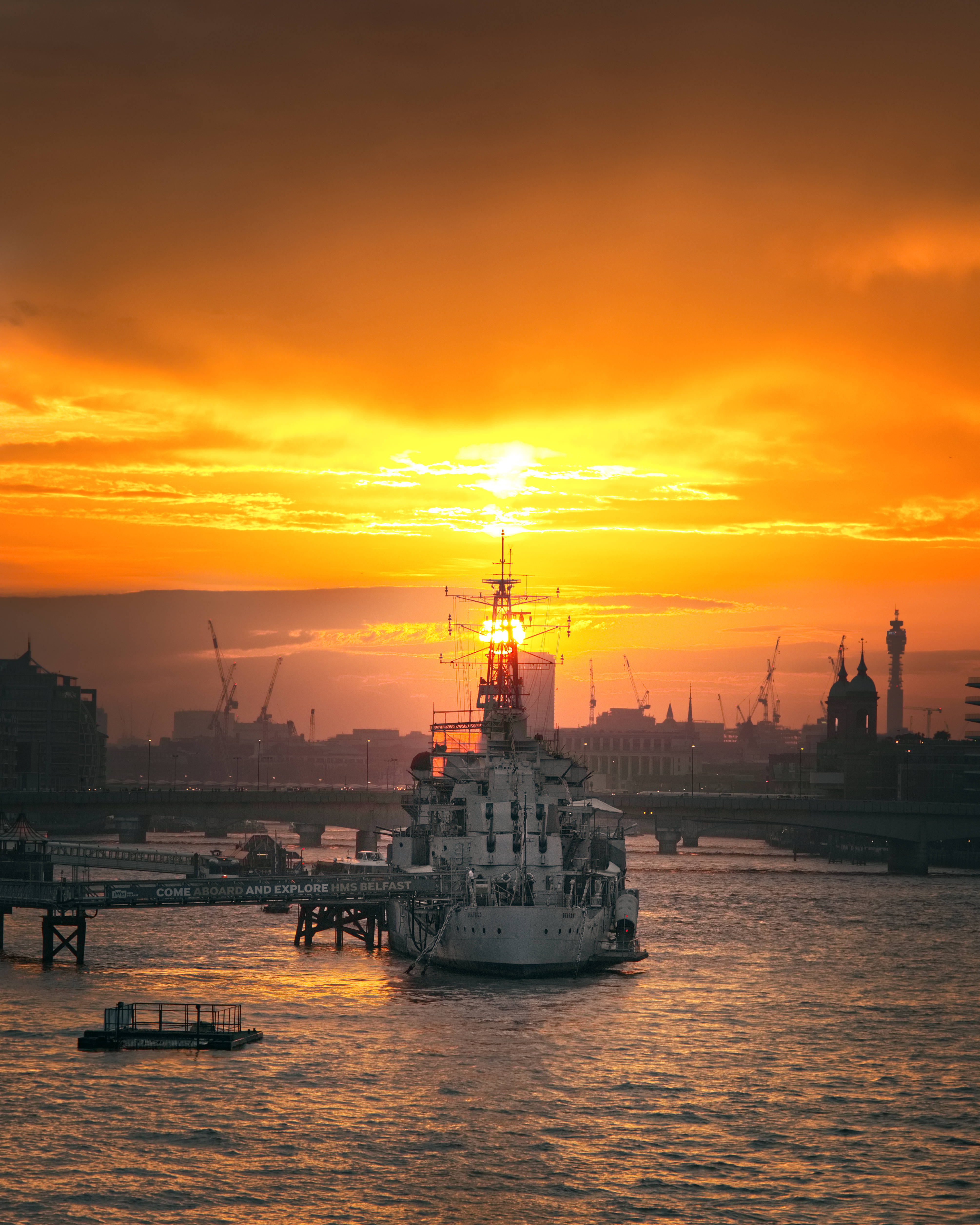 Warship 桌布 壁紙 — 一艘歷史戰艦在火橙色夕陽下剪影，背景是城市海濱，遠處有橋樑和起重機。