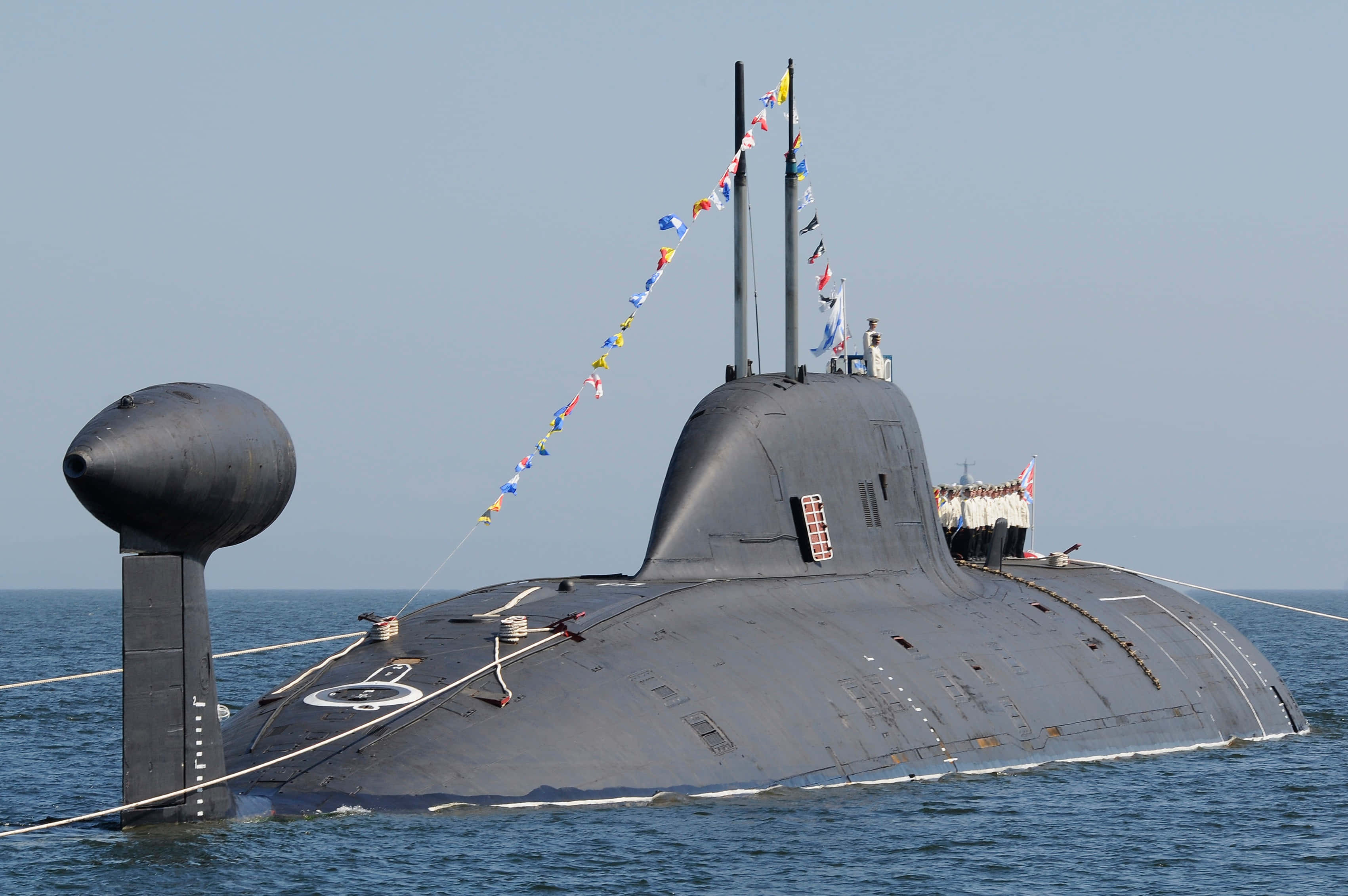 Submarine 桌布 壁紙 — 一艘流線型灰色潛艇在平靜的藍色海水中浮出水面，船員站在甲板上，桅杆上掛滿了彩色信號旗。