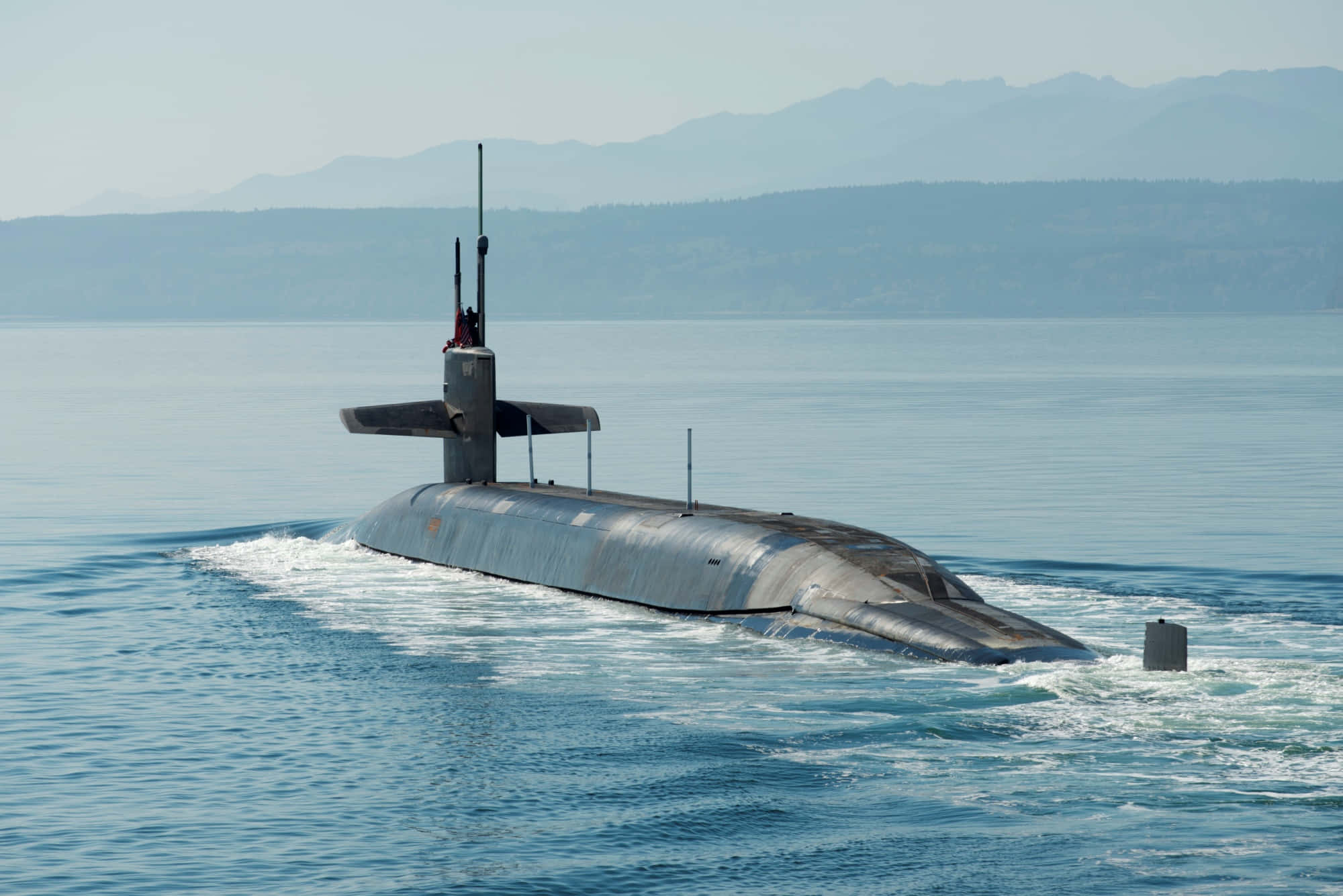 Submarine 桌布 壁紙 — 一艘流線型灰色潛艇在平靜的藍色海水中浮出水面，遠方有朦朧的山脈，天空晴朗。