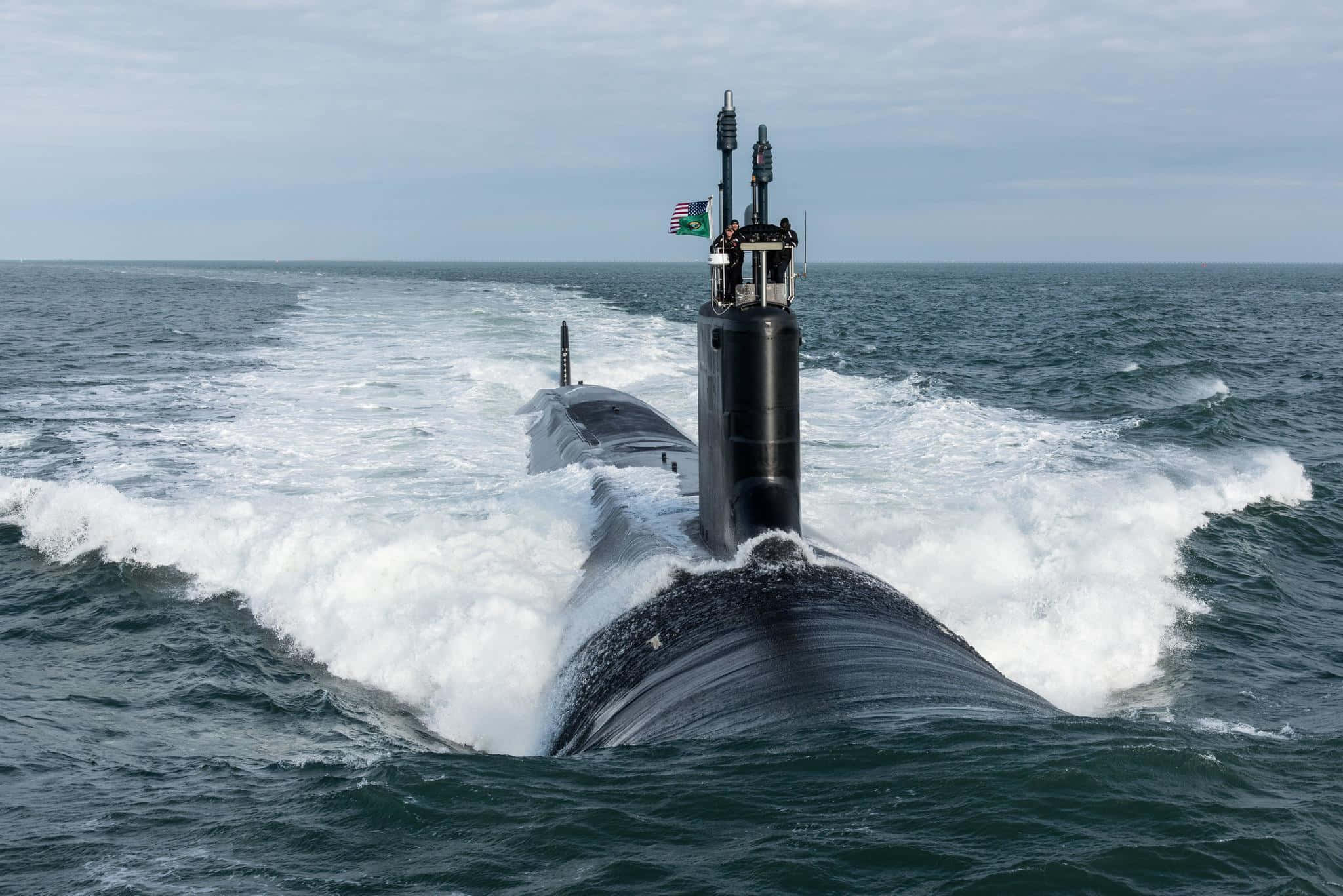 Submarine 桌布 壁紙 — 一艘流線型的黑色潛艇在波濤洶湧的海浪中穿行，在多雲的天空下留下泡沫般的白色尾跡。
