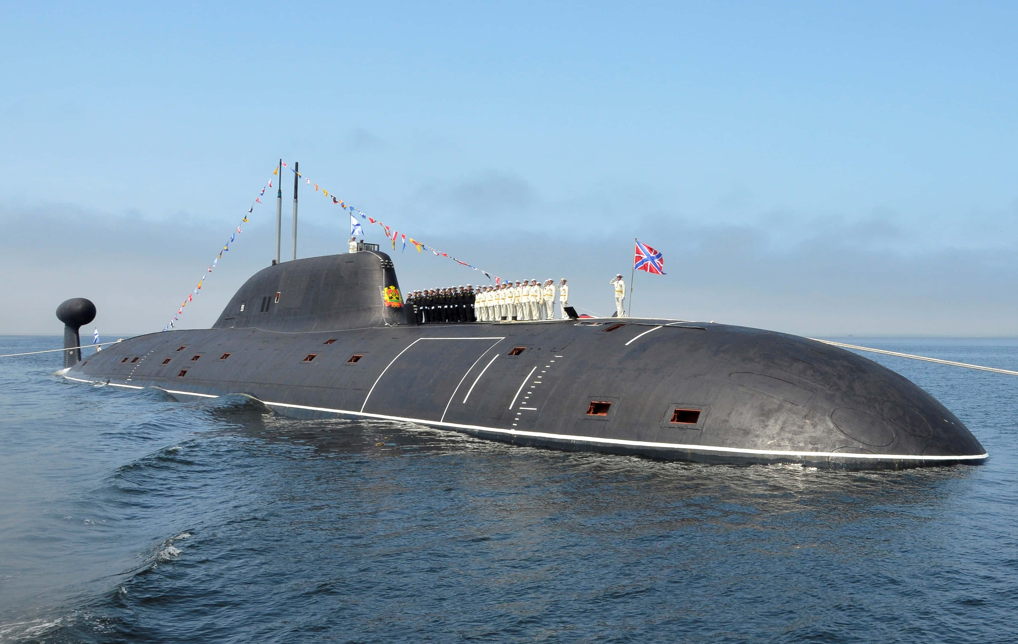 Submarine 桌布 壁紙 — 一艘流線型黑色潛艇在平靜的藍色海水中浮出水面，身穿白色制服的船員在晴朗的天空下於甲板上立正。