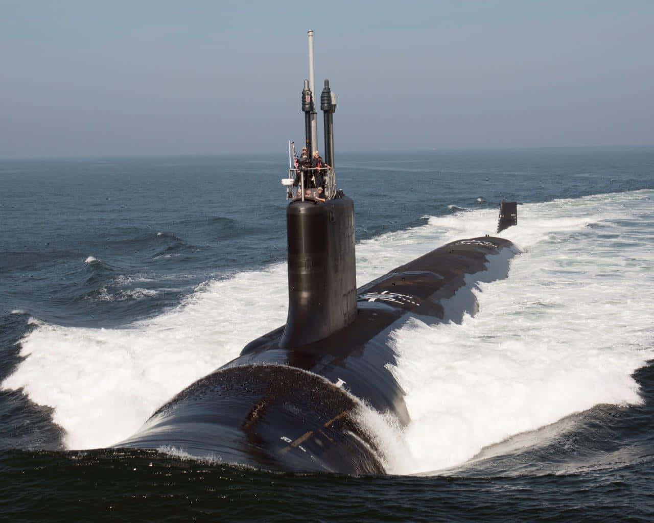Submarine 桌布 壁紙 — 一艘流線型黑色潛艇在波濤洶湧的藍色海水中浮出水面，在朦朧的天空下留下白色泡沫尾跡。