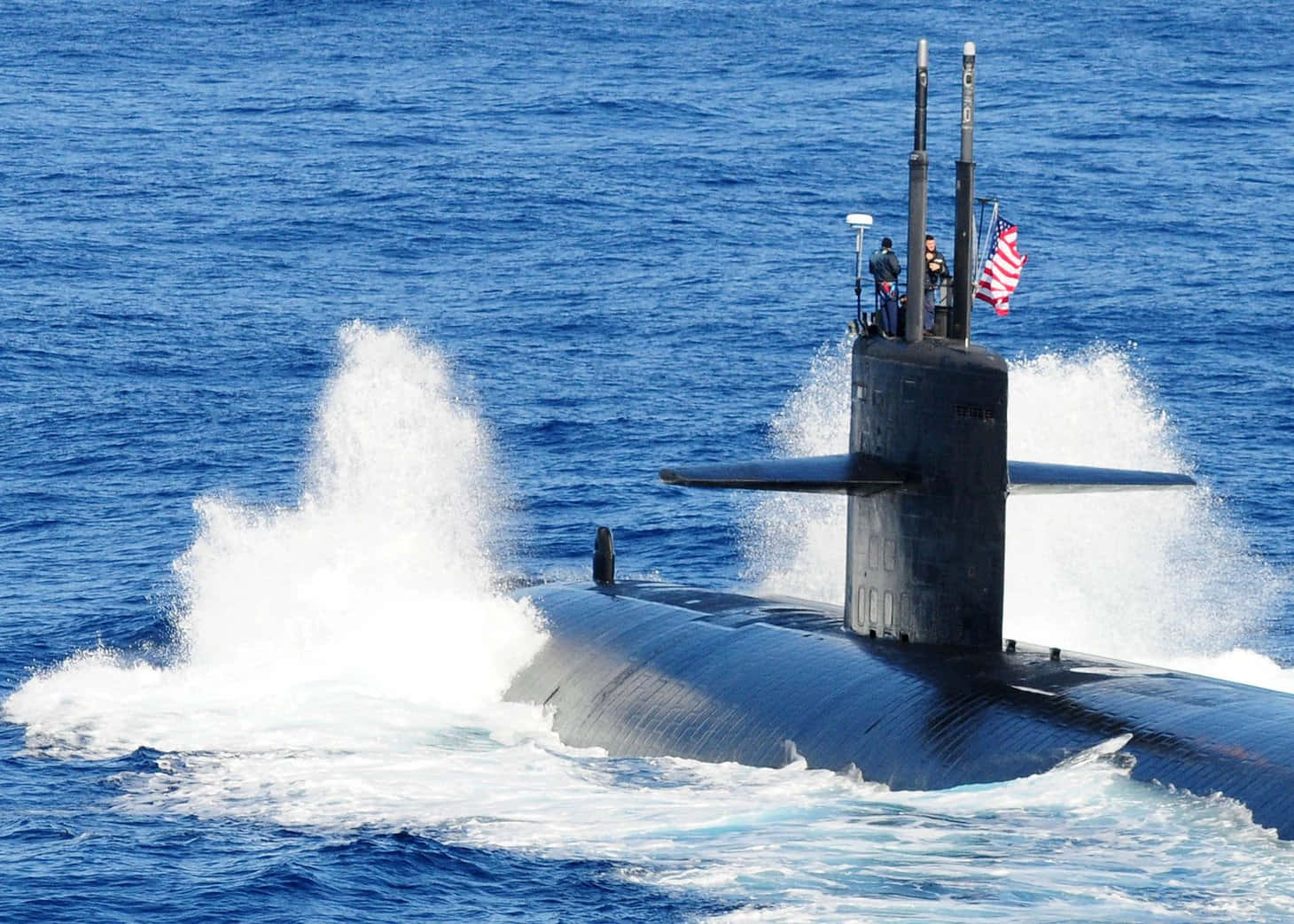 Submarine 桌布 壁紙 — 一艘流線型黑色潛艇從深藍色海水中浮出，白色水花在其指揮塔周圍飛濺，船員站立其上，美國國旗飄揚。