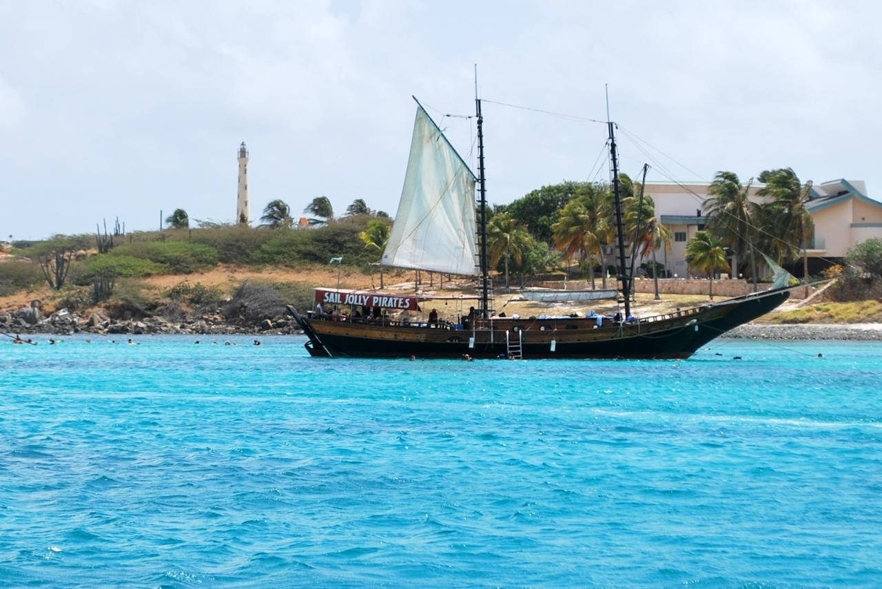 Aruba 桌布 壁紙 — 一艘海盜主題帆船在燈塔和棕櫚樹環繞的海岸附近的碧綠海水中航行。