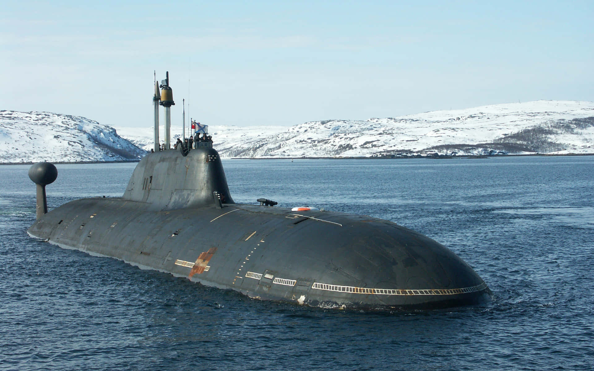 Submarine 桌布 壁紙 — 一艘深灰色潛艇在平靜的藍色水域中浮出水面，背景是白雪覆蓋的山脈和晴朗的天空。
