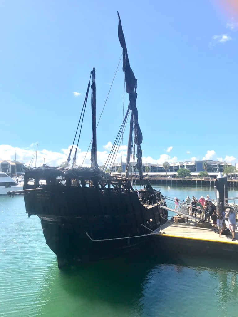 Pirate 桌布 壁紙 — 一艘深色海盜船掛著破爛的帆，停泊在陽光明媚的碼頭，遊客聚集在棧橋上。