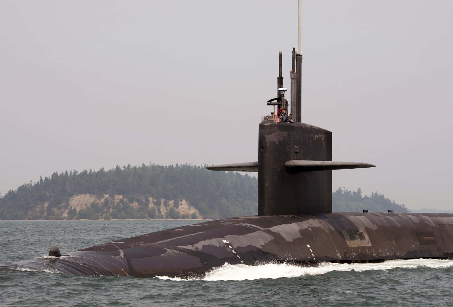 Submarine 桌布 壁紙 — 一艘深色軍用潛艇在平靜水域中浮出水面，背景朦朧中有一座樹木茂密的島嶼。
