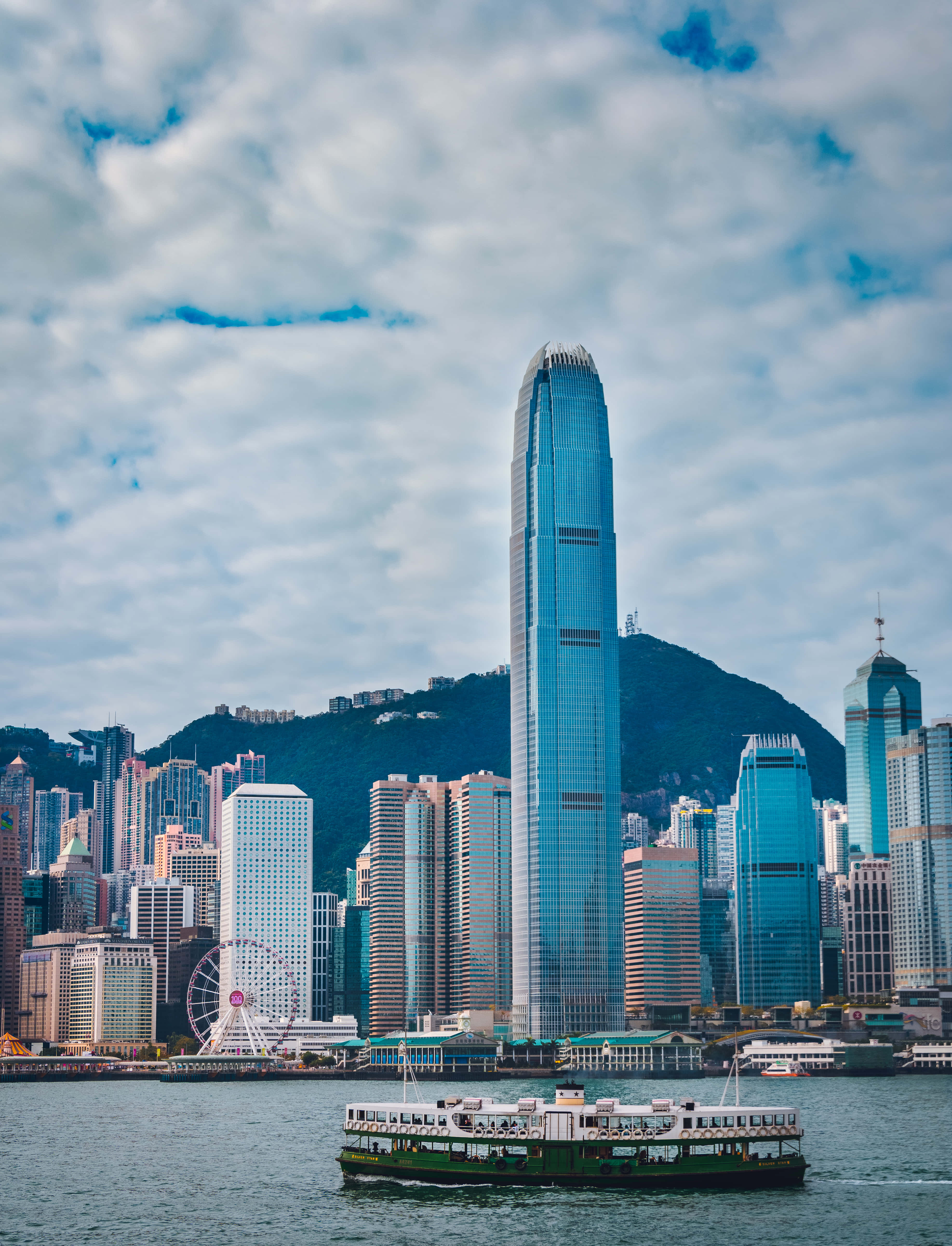香港 桌布 壁紙 — 一艘渡輪在香港港口滑行，下方是高聳的玻璃摩天大樓和多雲的天空。
