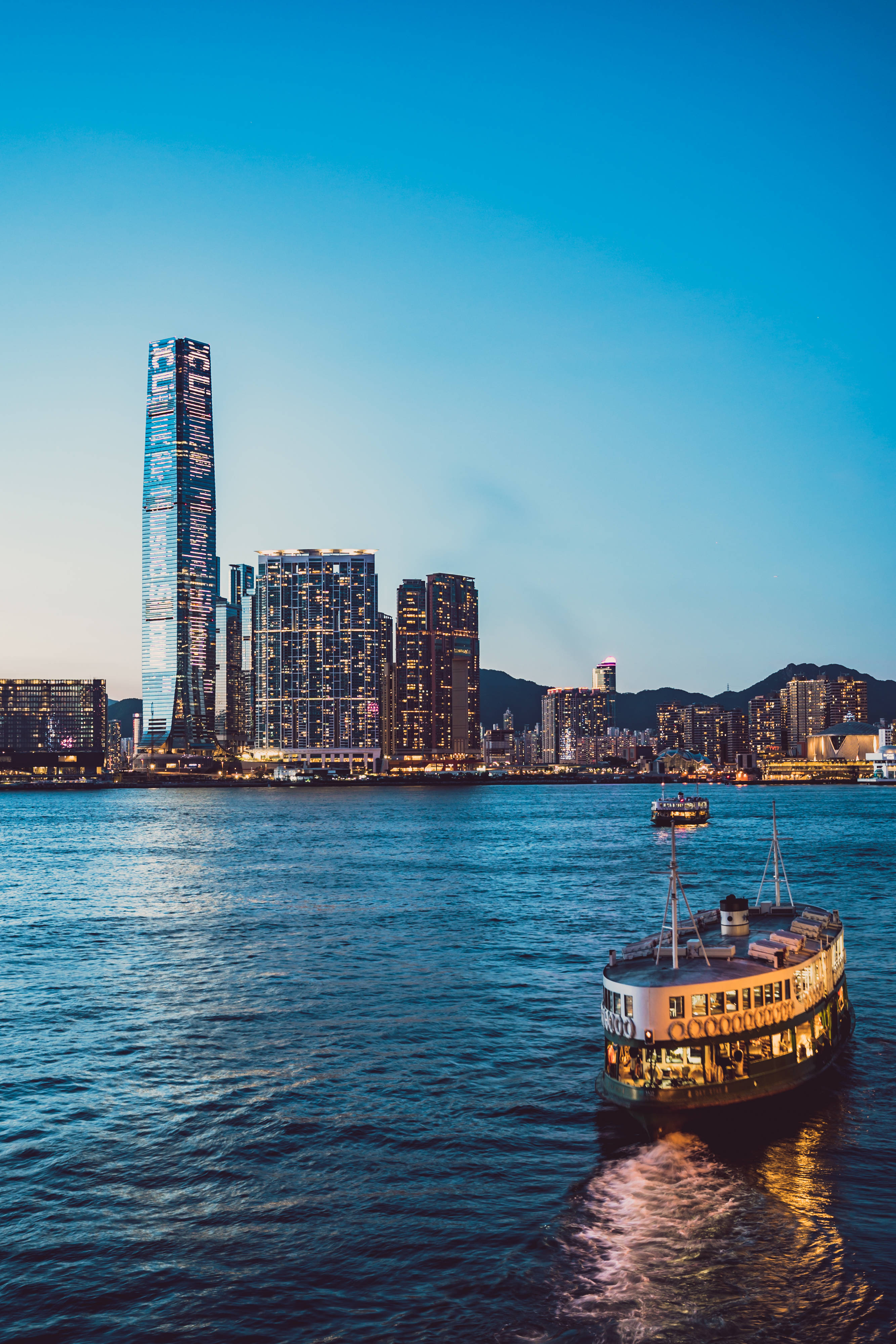 香港 桌布 壁紙 — 一艘渡輪在黃昏時分滑過平靜的港口，背後是閃爍著高聳摩天大樓的城市天際線。