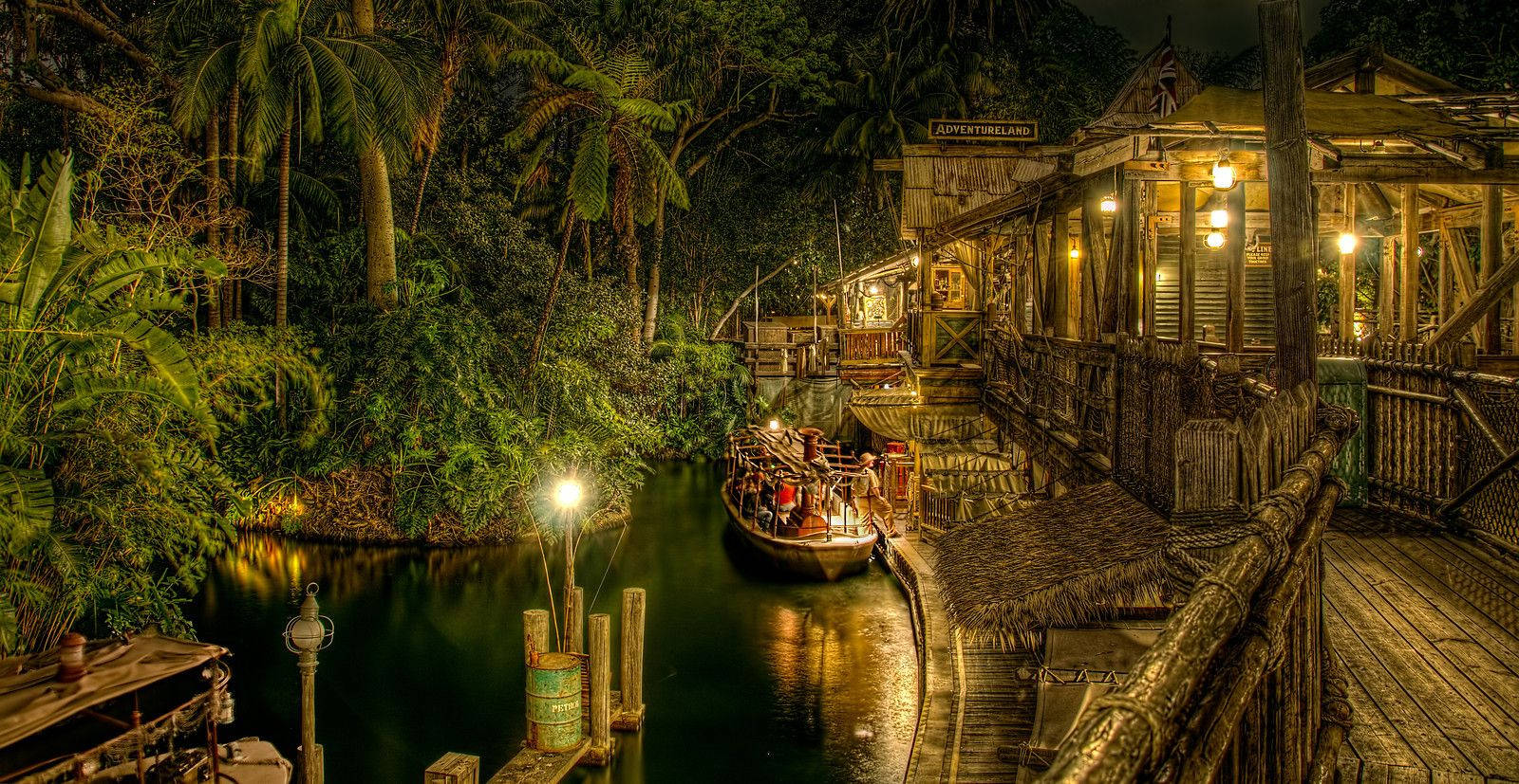 Jungle Cruise 桌布 壁紙 — 一艘溫暖照明的木船在夜間停泊在茂密熱帶植被下的鄉村河畔車站。