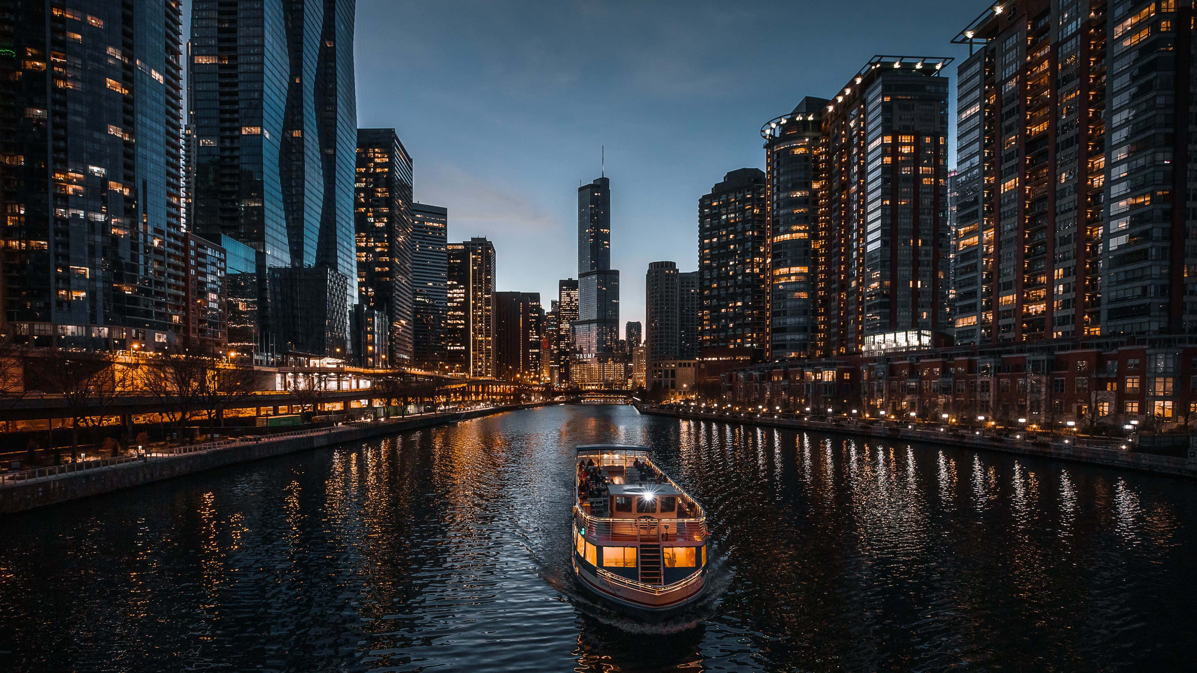 4K Ultra HD Chicago 桌布 壁紙 — 一艘溫暖照明的河船滑過城市運河，兩旁是黃昏時發光的摩天大樓。