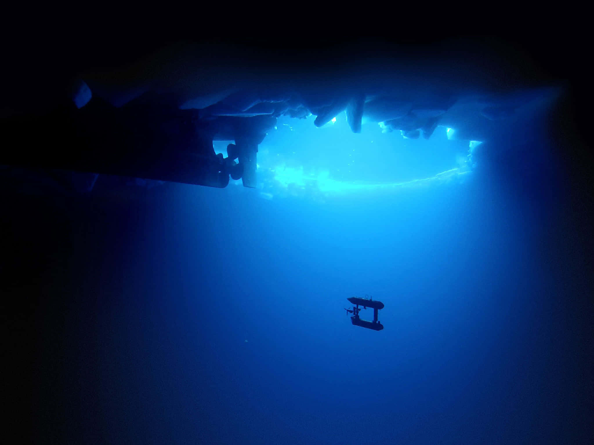 Scary Underwater 桌布 壁紙 — 一艘潛水器在遠方藍光照亮的冰洞下，微弱地發著光，緩緩降入深淵。
