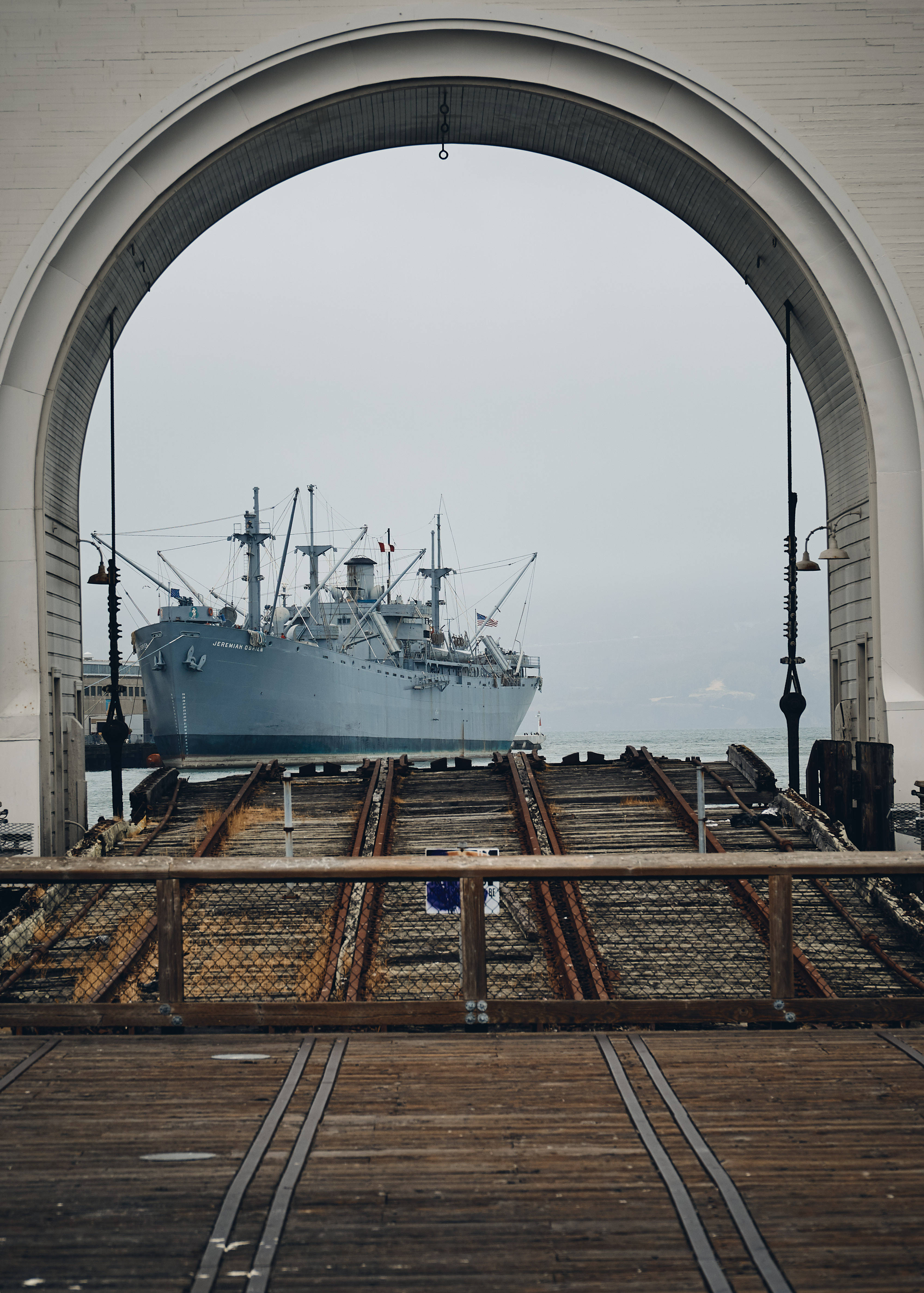 Warship 桌布 壁紙 — 一艘灰色大船停泊在經過風化的木製斜坡之外，斜坡由白色拱形結構框住，天空多雲。