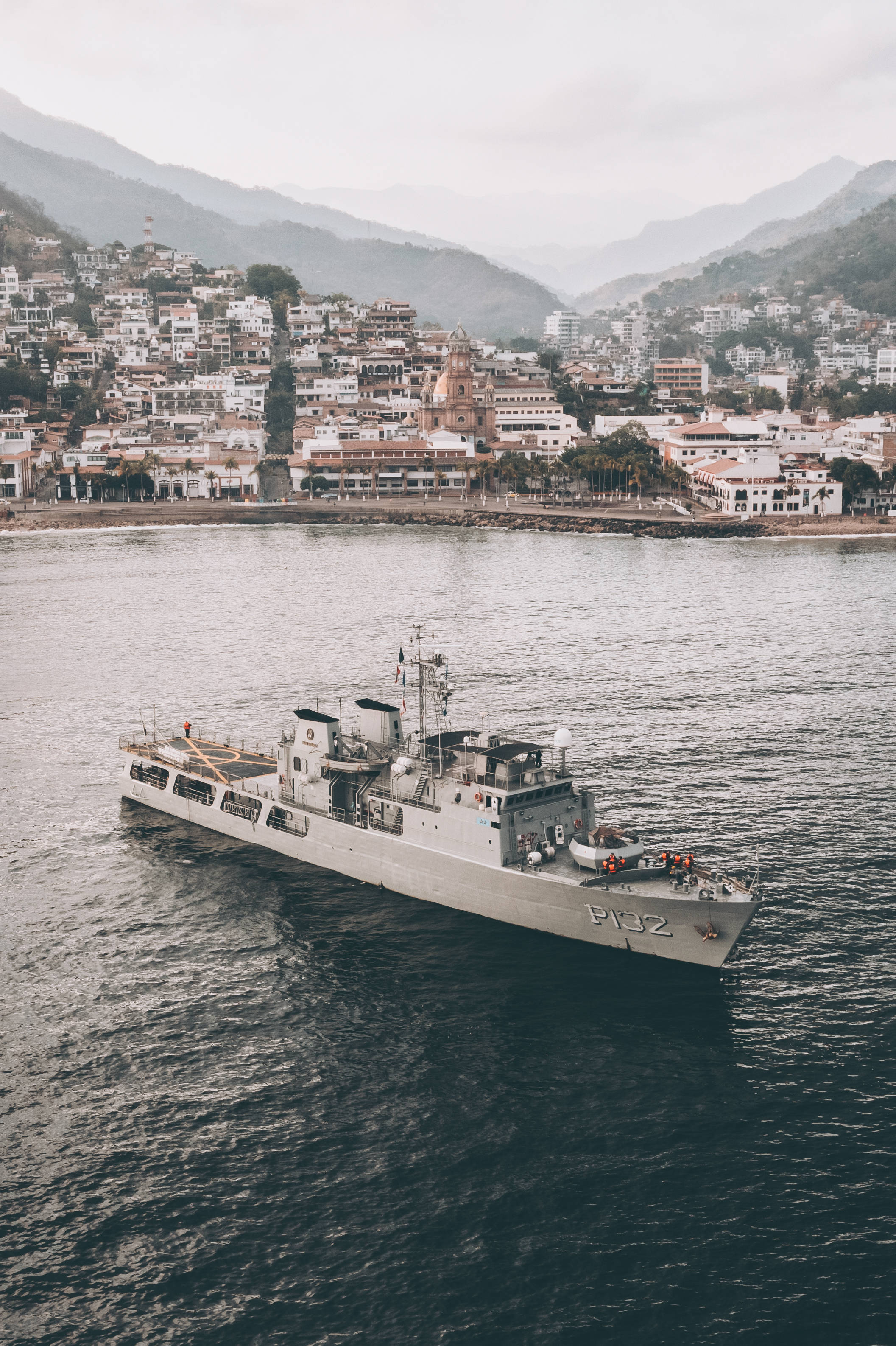 Warship 桌布 壁紙 — 一艘灰色海軍艦艇在黑暗水域中平靜航行，背景是沿海小鎮和霧氣繚繞的山脈。