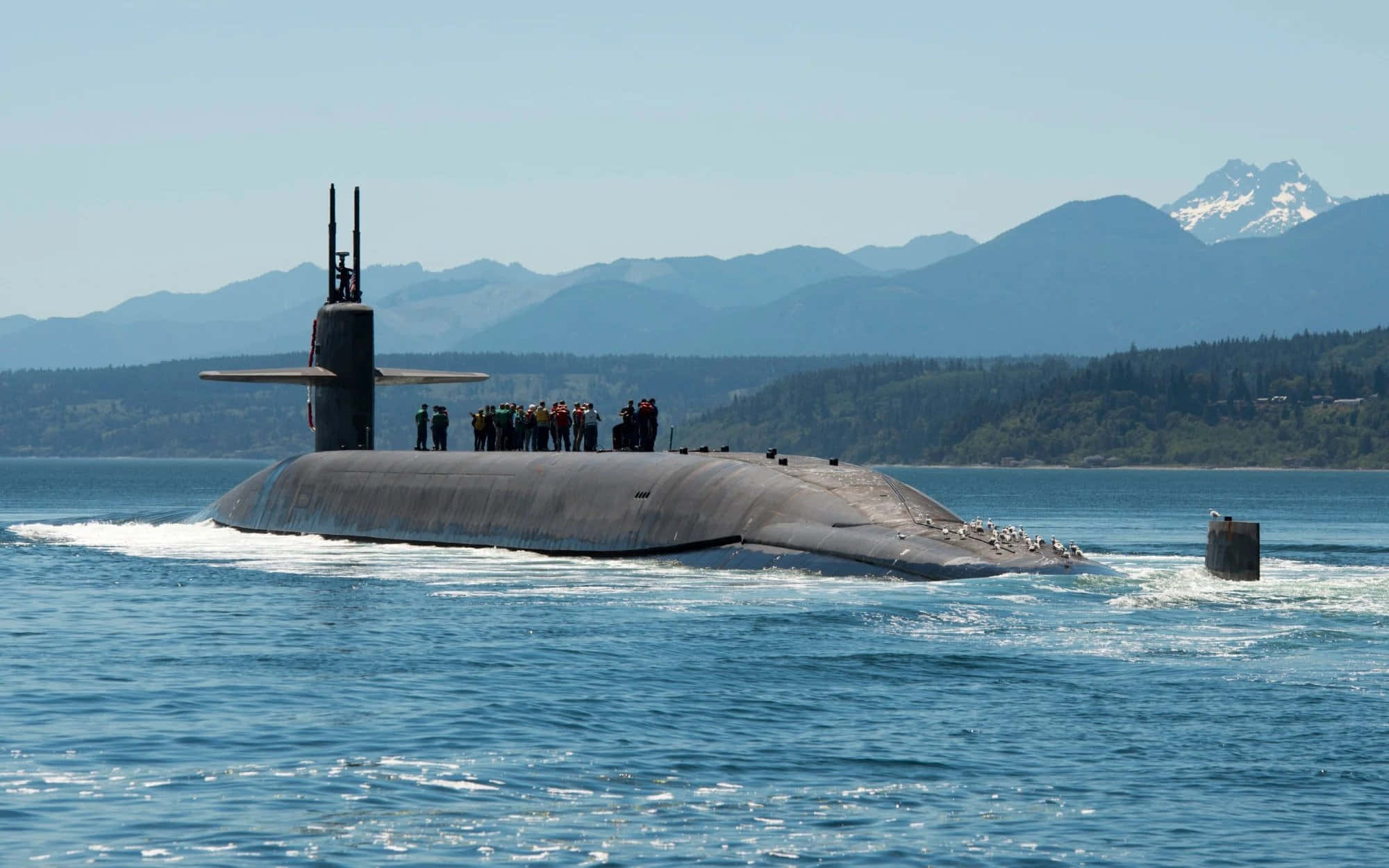 Submarine 桌布 壁紙 — 一艘灰色潛艇在藍色海水中浮出水面，甲板上有船員，遠處有積雪覆蓋的山脈，天空晴朗。