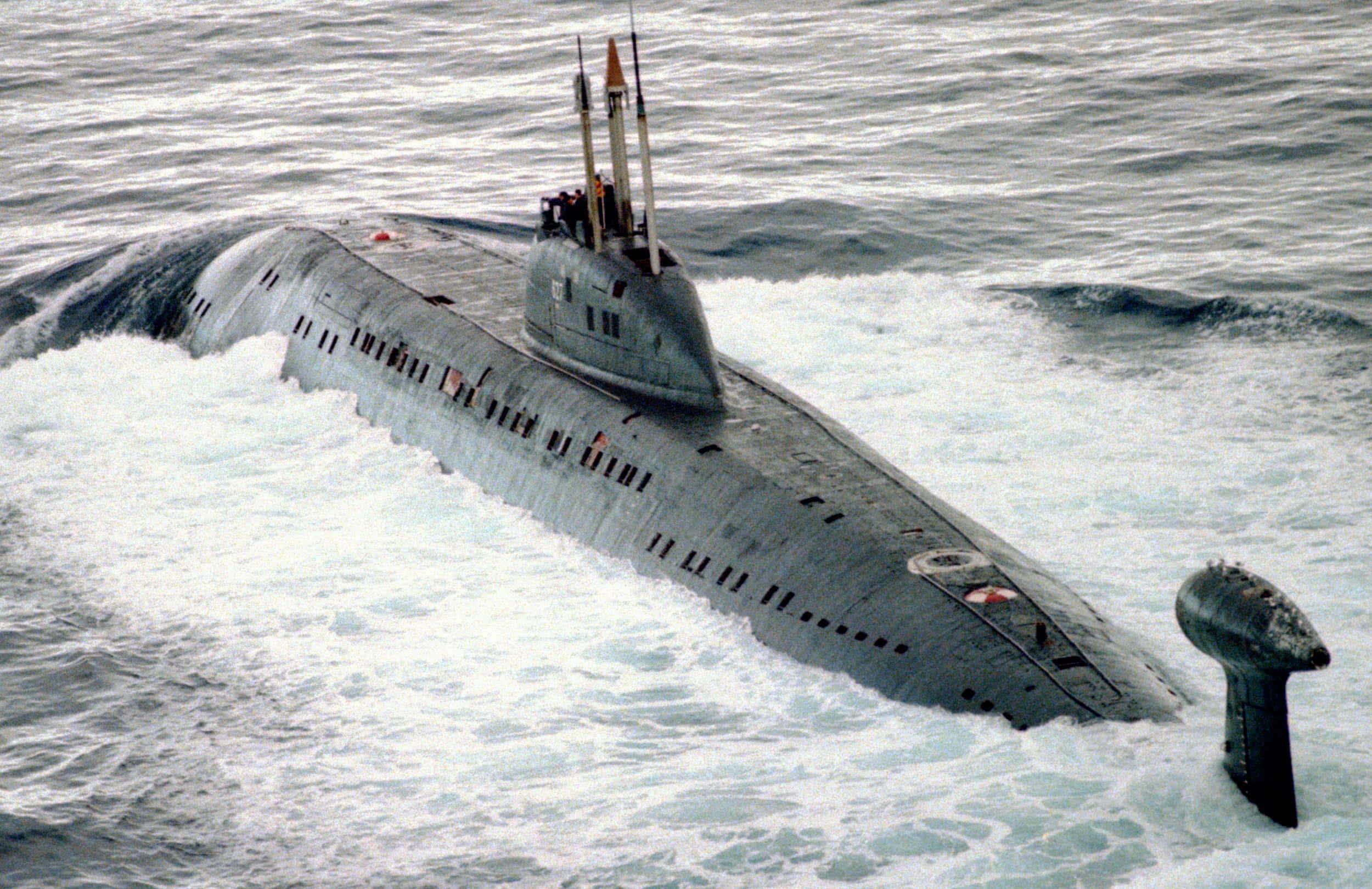 Submarine 桌布 壁紙 — 一艘灰色軍用潛艇在波濤洶湧的海浪中浮出水面，潛望鏡和飛彈發射管清晰可見。