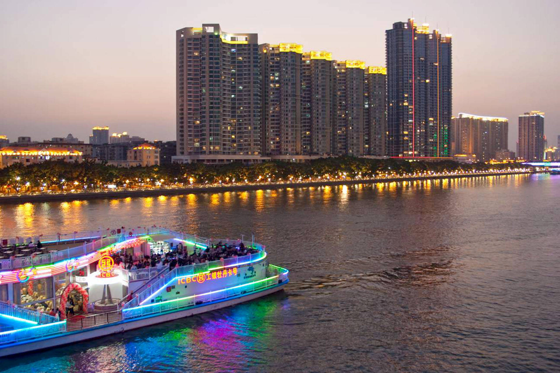 Guangzhou 桌布 壁紙 — 一艘燈火通明、帶有彩色霓虹燈的河上遊船，在黃昏時分滑過倒映在水中的高聳摩天大樓。