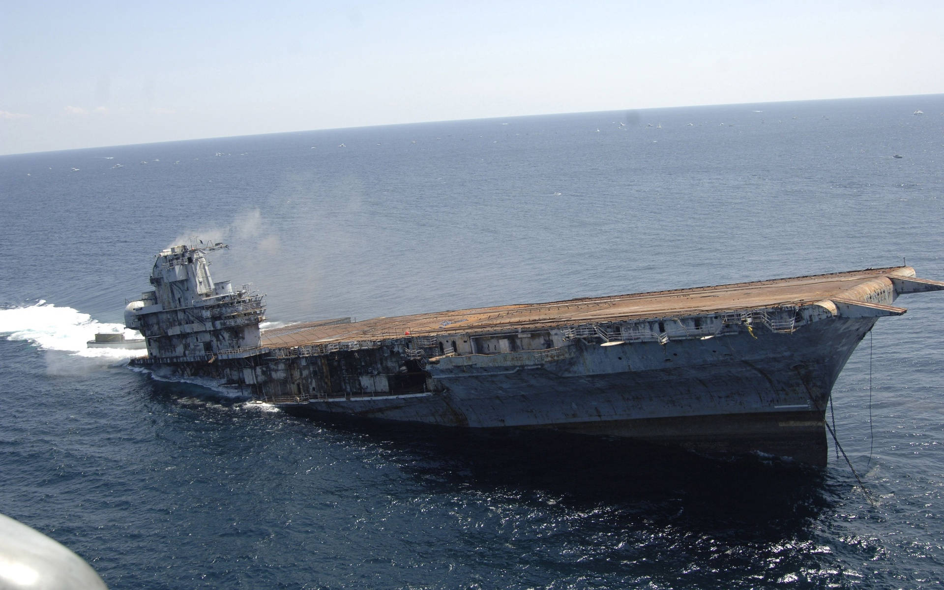 Warship 桌布 壁紙 — 一艘生鏽的航空母艦在清澈的天空下傾斜在深藍色的海洋中，甲板上冒著煙。