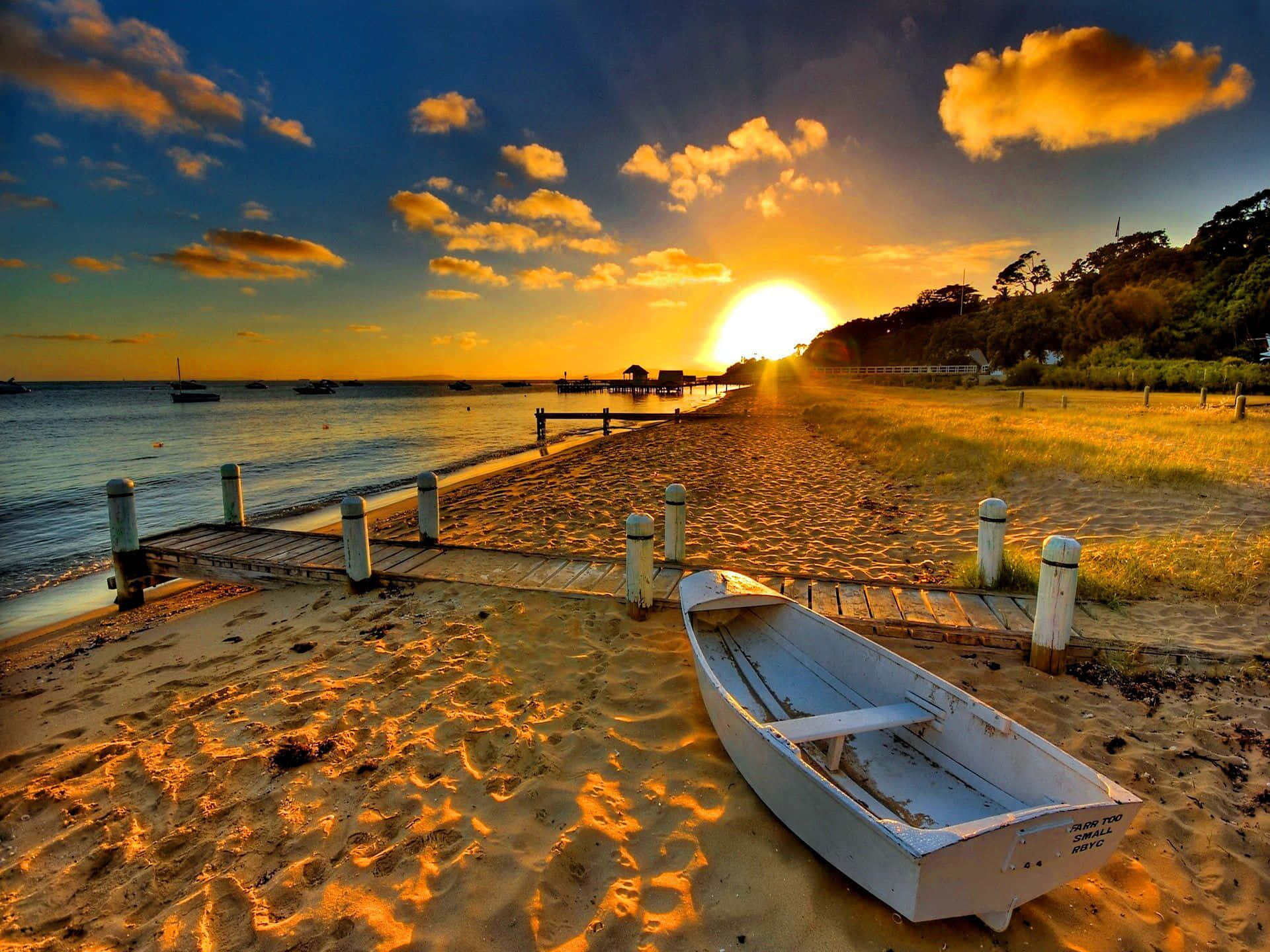 Beach Sunset HD 桌布 壁紙 — 一艘白色划艇在日落時分停靠在金色沙灘上，沐浴在溫暖的光線中，太陽在碼頭和遠處船隻後方緩緩落下。