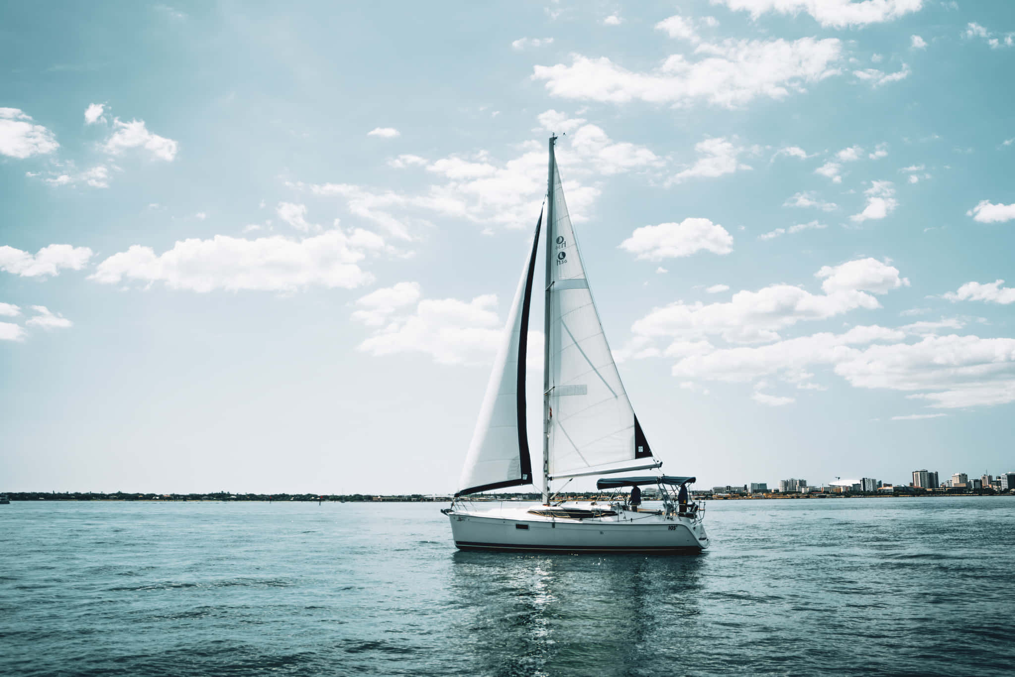 Sailboat 桌布 壁紙 — 一艘白色帆船在晴朗的天空下，於平靜的藍色水面上滑行，天空點綴著蓬鬆的雲朵。