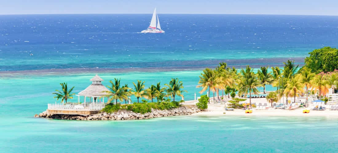 Jamaican Island 桌布 壁紙 — 一艘白色帆船在熱帶島嶼附近碧綠的海水中滑行，島上有棕櫚樹和茅草涼亭。