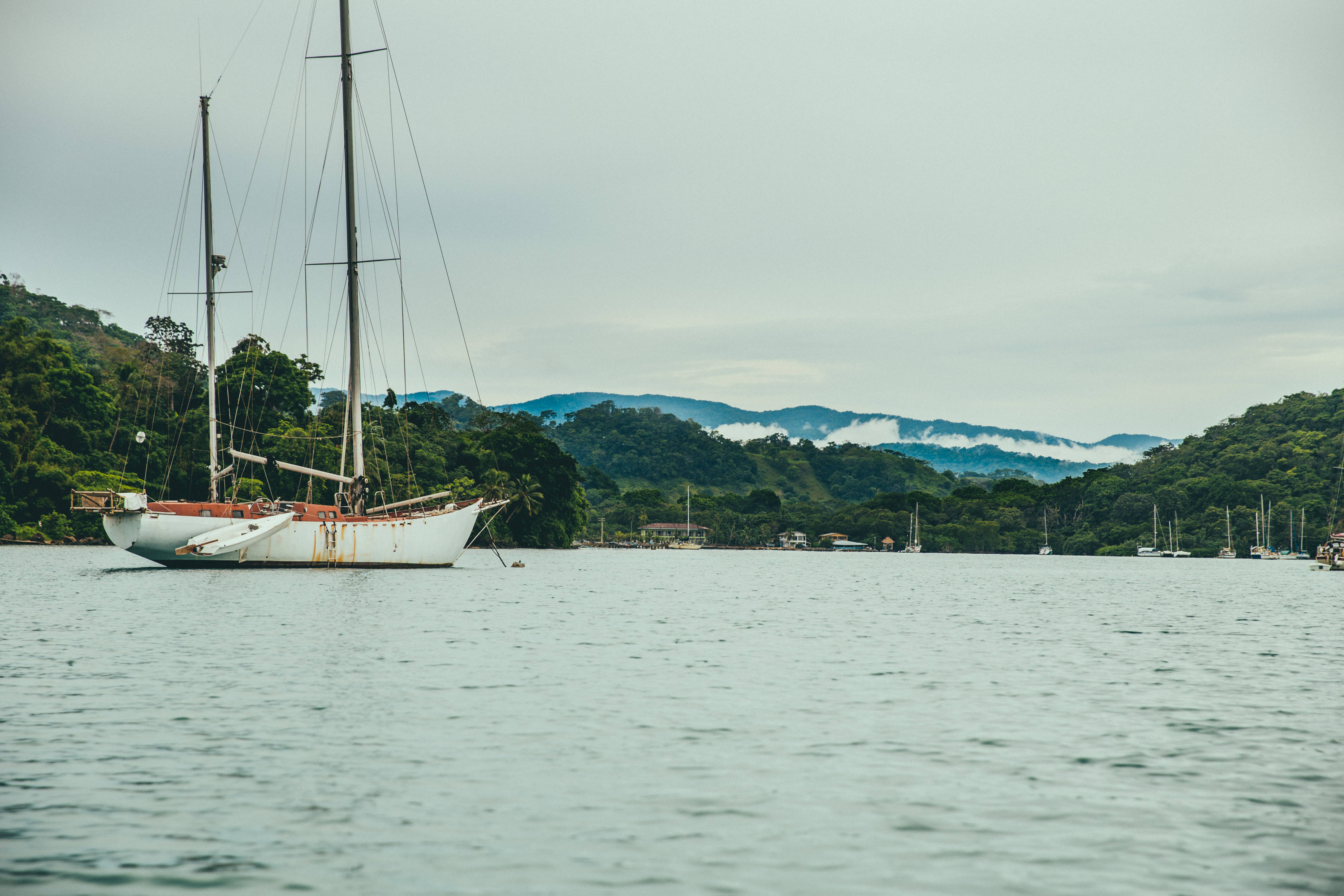 Panama 桌布 壁紙 — 一艘白色帆船在陰天之下，平靜地停泊在寧靜的水面上，周圍是茂密的綠色山丘和遠處霧濛濛的山脈。