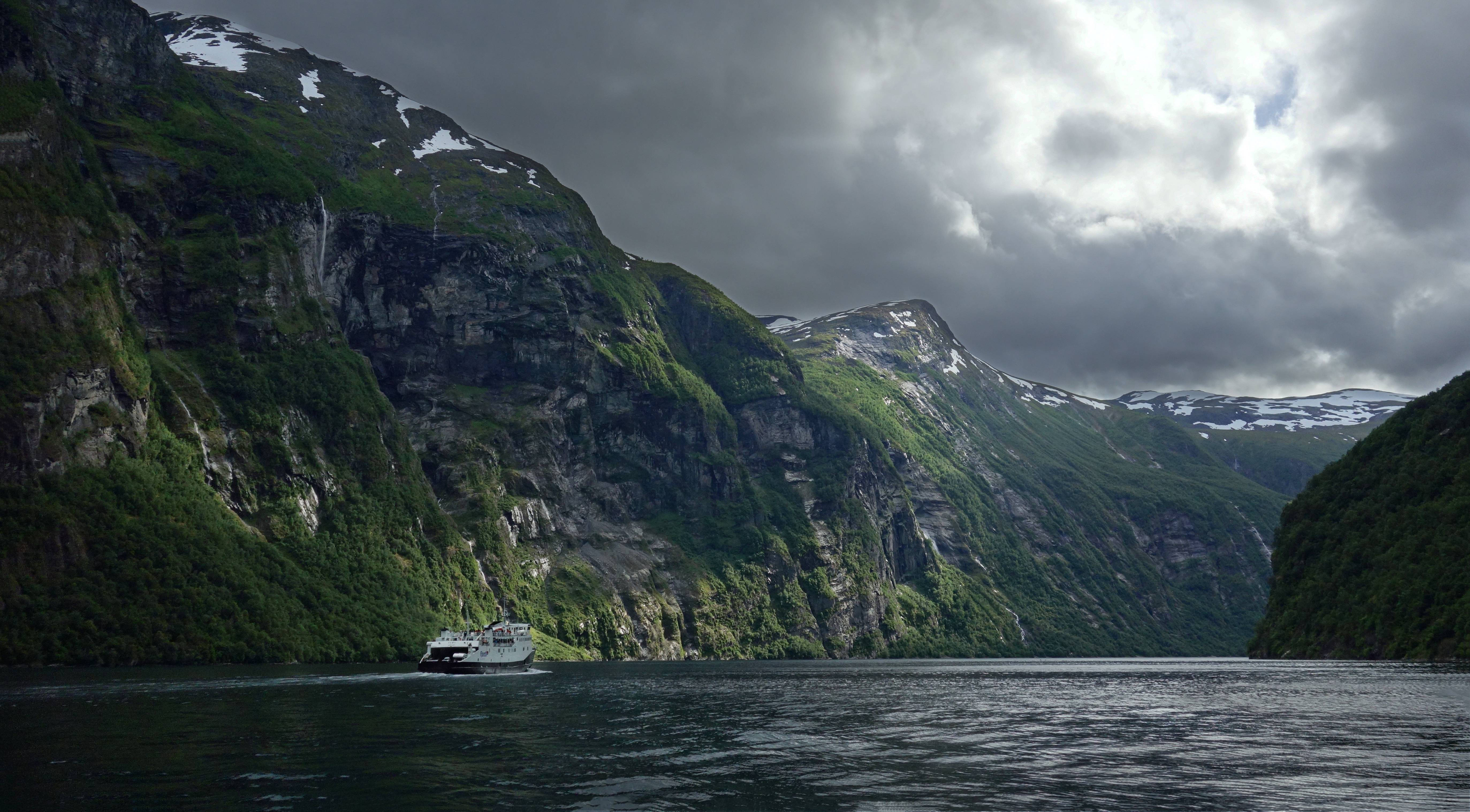 Norway 桌布 壁紙 — 一艘白色渡輪在戲劇性的多雲天空下，航行在兩旁高聳的綠色山脈（山頂積雪）之間的狹窄峽灣中。