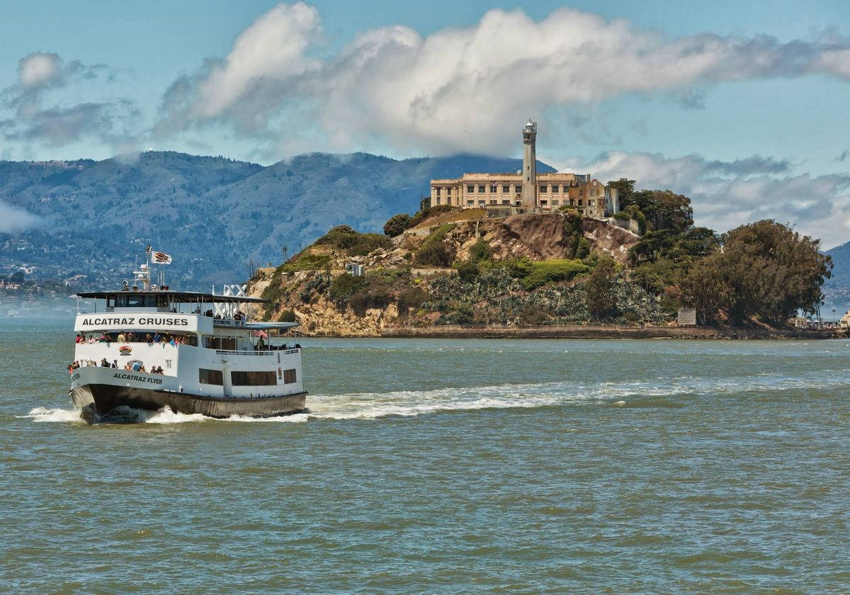 美國 桌布 壁紙 — 一艘白色的 Alcatraz Cruises 渡輪在部分多雲的藍天下駛過歷史悠久的島嶼監獄。