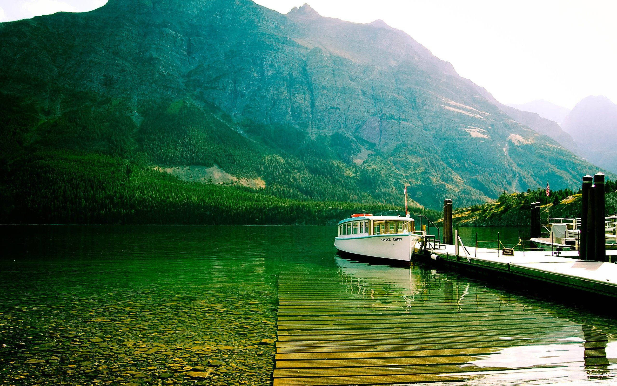Lake 桌布 壁紙 — 一艘白色觀光船停泊在木製碼頭，湖面如鏡，碧綠清澈，四周被高聳的森林覆蓋的山脈環繞，陽光普照。