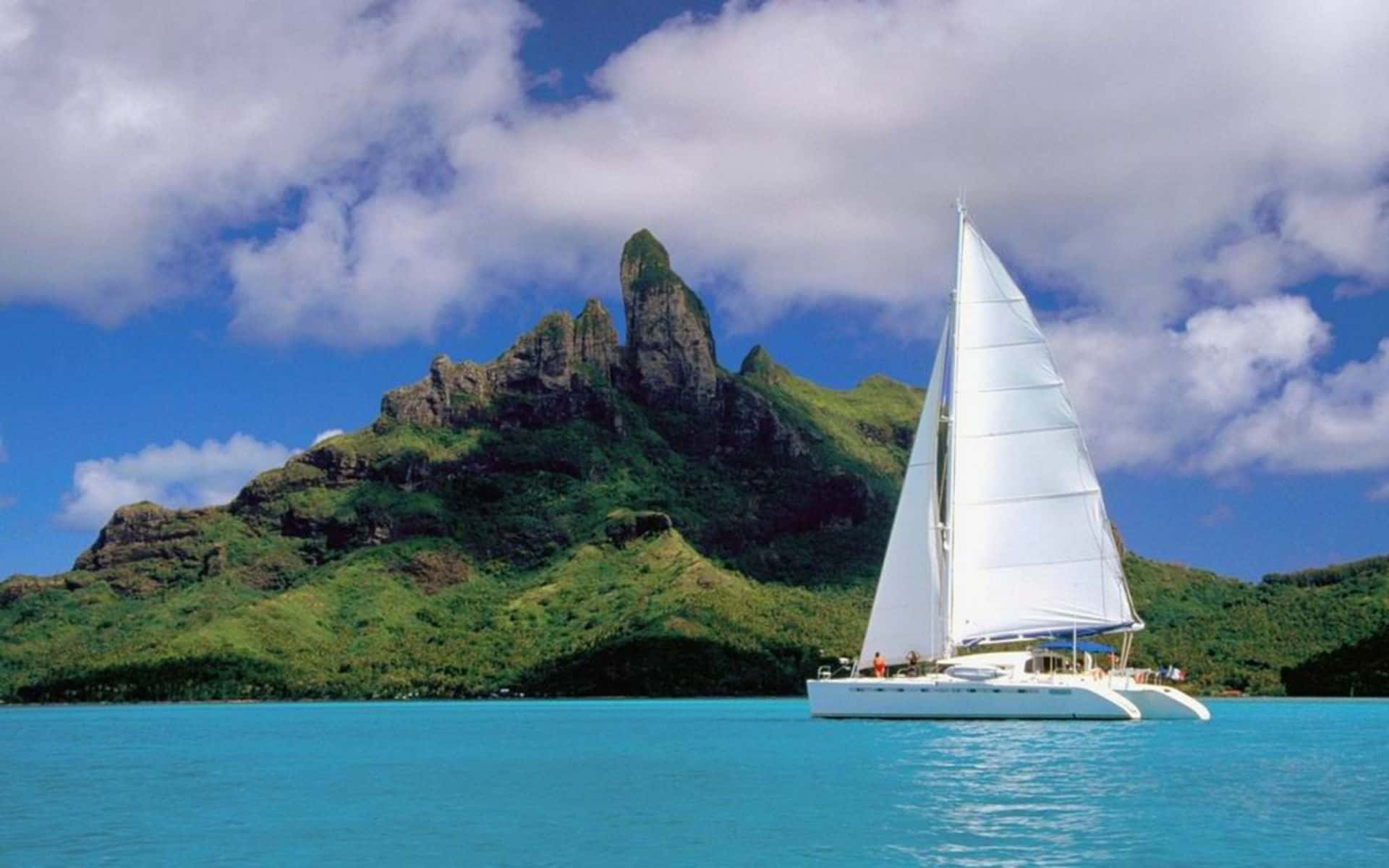 Sailboat 桌布 壁紙 — 一艘白色雙體船在碧綠的海水上優雅航行，背後是鬱鬱蔥蔥的綠色山脈，在明亮的藍天下戲劇性地聳立。